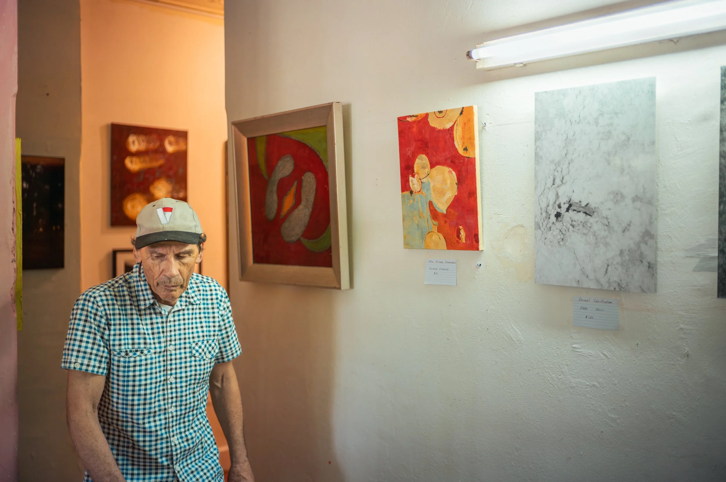 An elderly man in a checkered shirt and beige cap standing in an art gallery, looking down, with colorful abstract paintings on the wall behind him.