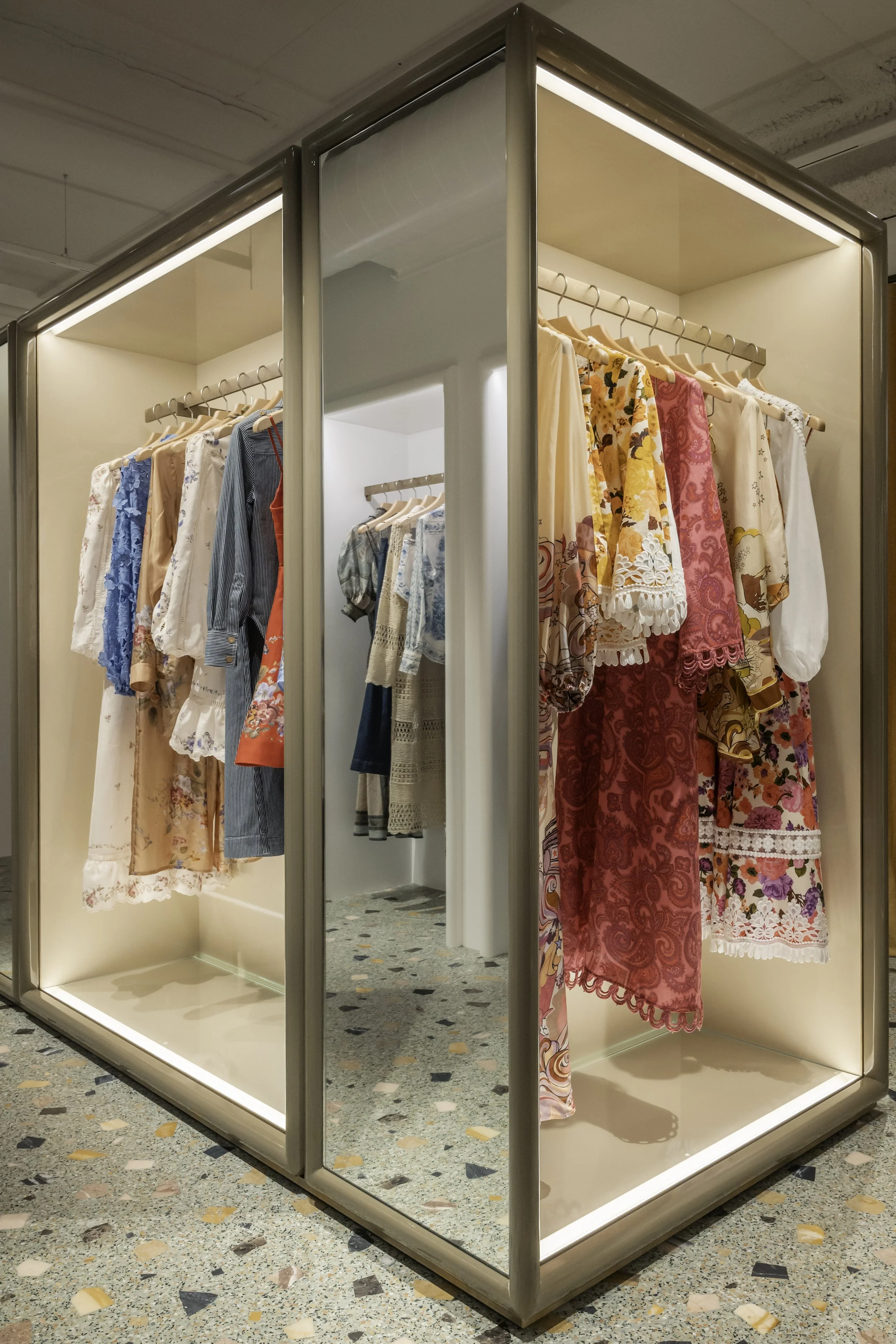 Clothing display with dresses and tops in a store, enclosed in glass cases with lighting.