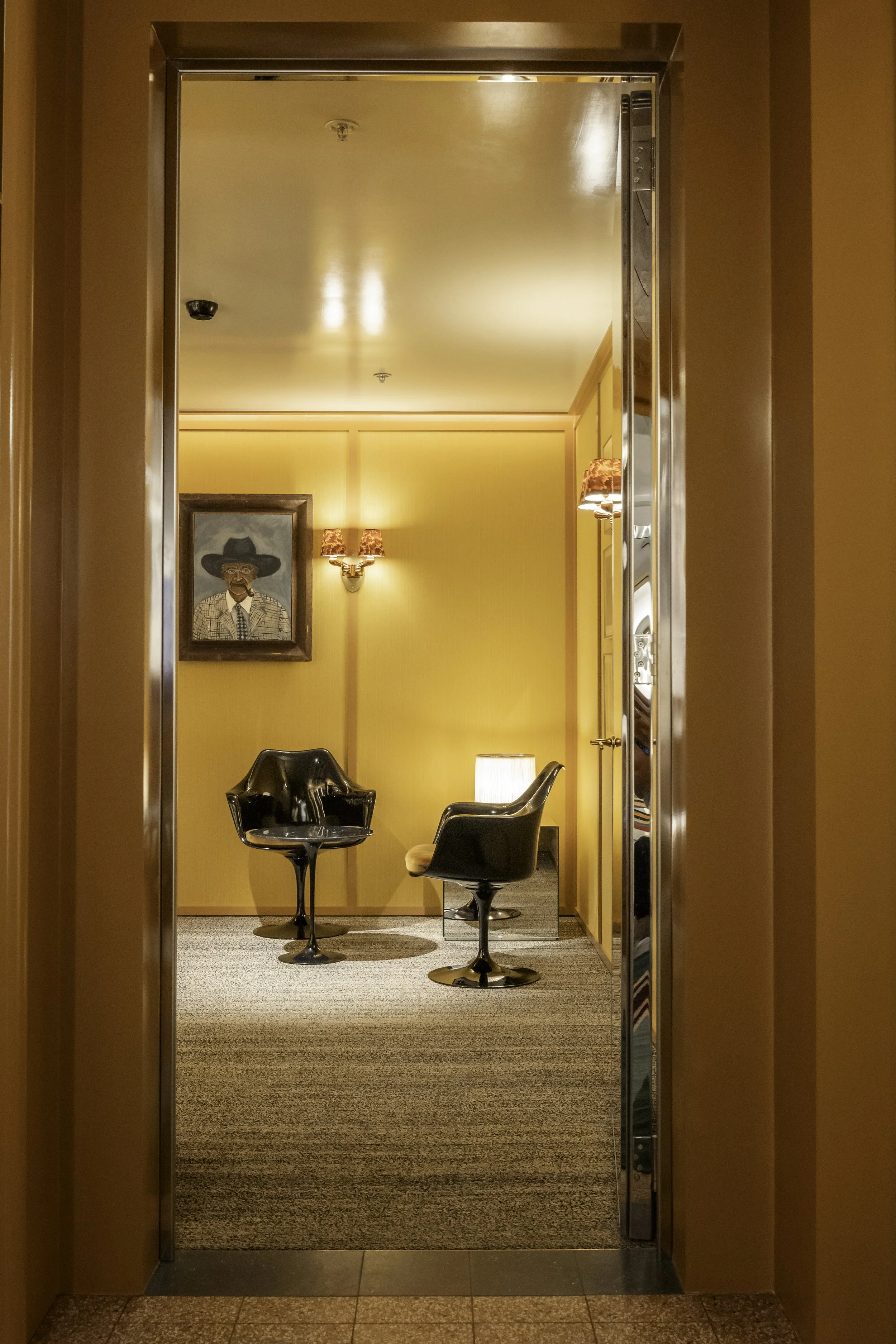 View through an open doorway into a cozy sitting area with two black chairs, a small table with a lamp, yellow walls, a framed painting of a man with a hat, and wall-mounted lamps.