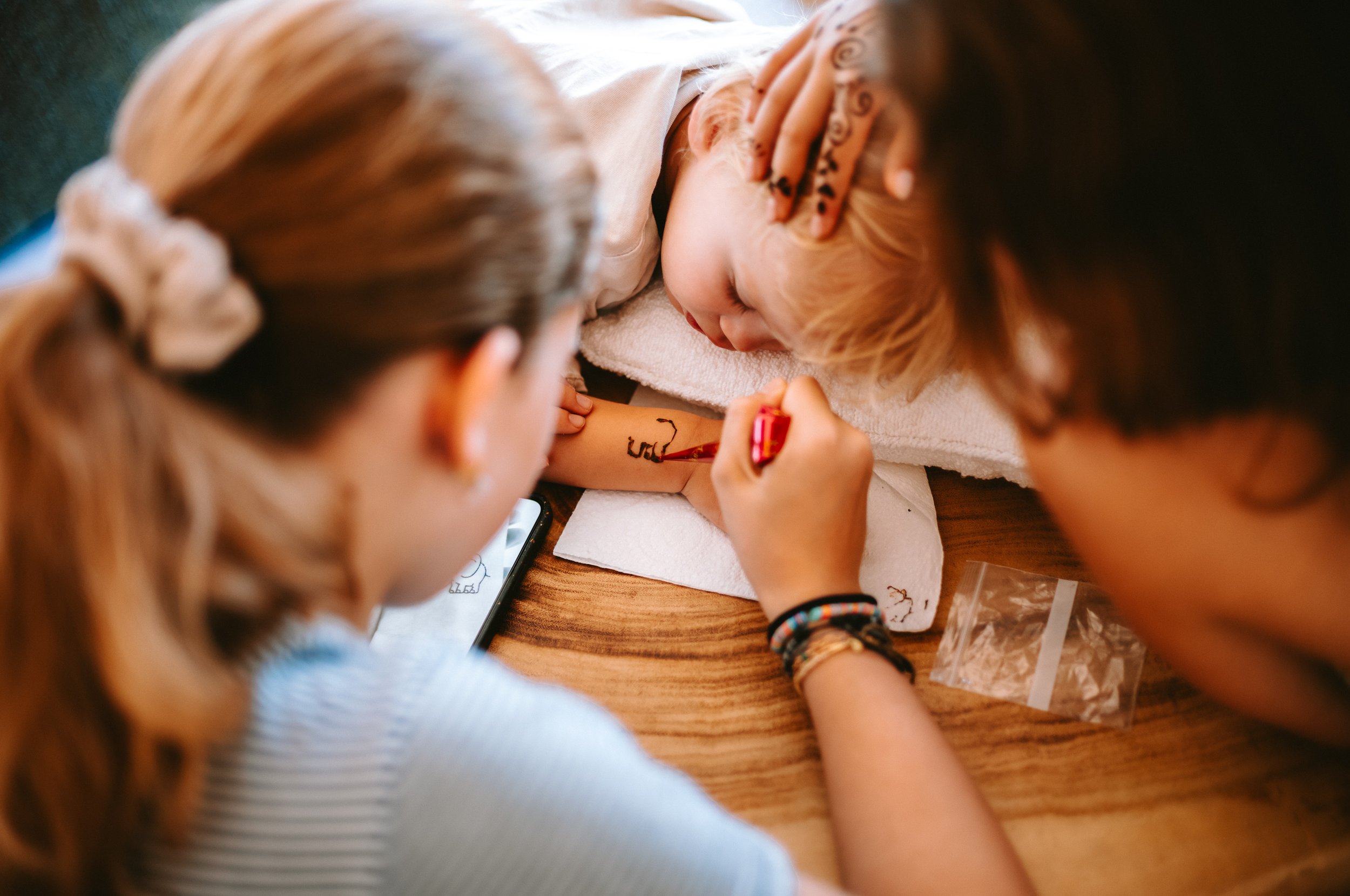 A young girl is lying on a towel having her arm painted with a temporary tattoo by a teenage girl. The teenage girl is holding a red marker and is focused on creating the tattoo.