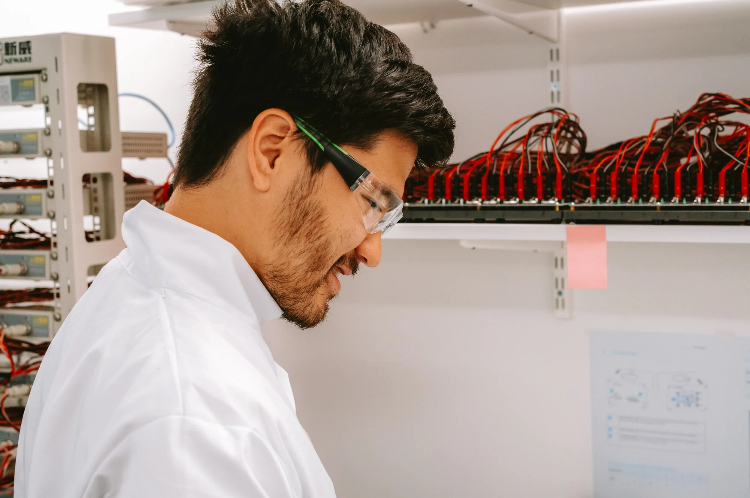 A man wearing safety goggles working on electronic components in a lab or data center with server racks and interconnected red cables in the background.