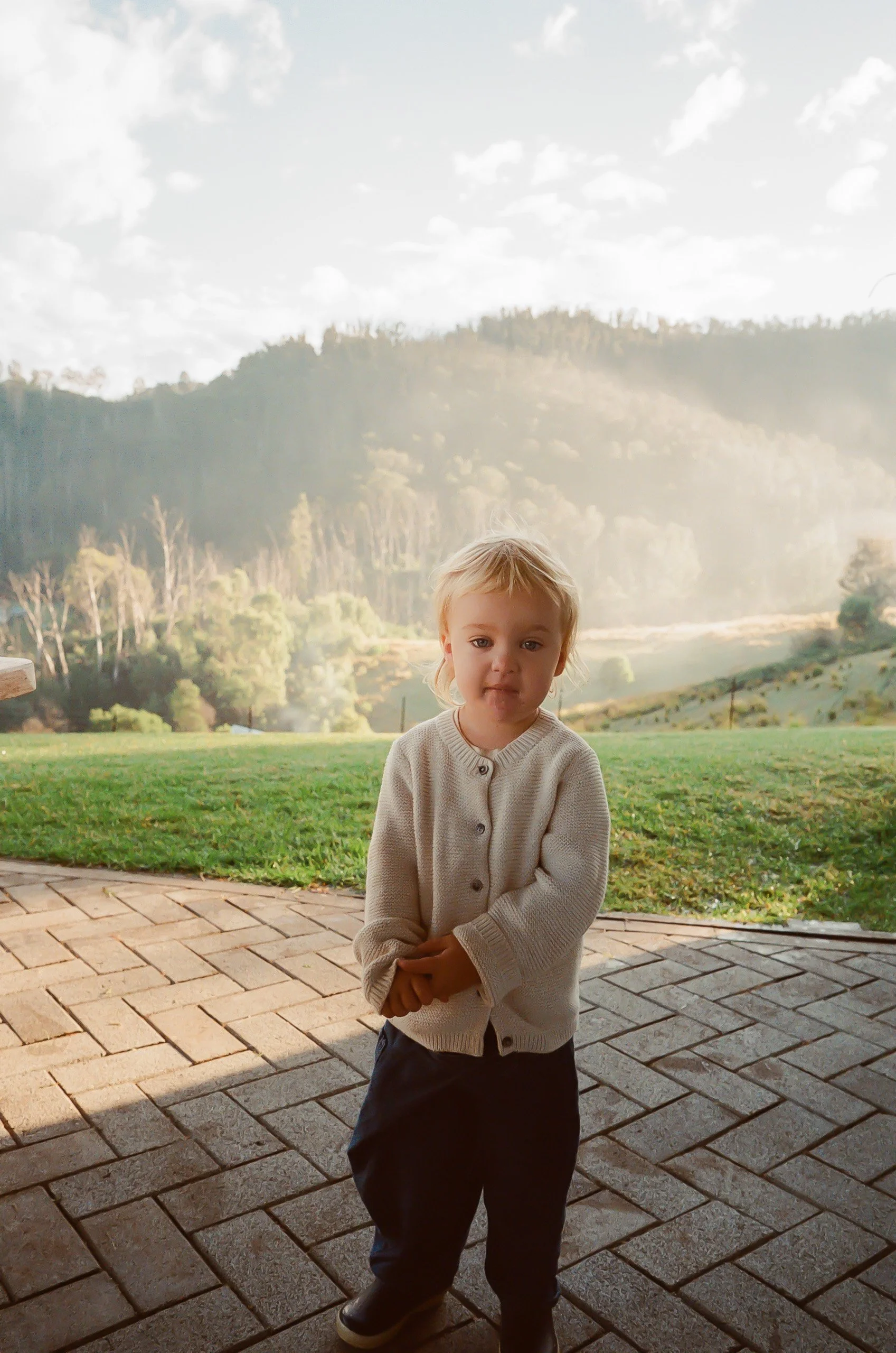 A young child with blonde hair wearing a beige cardigan and dark pants standing outdoors on a brick path, with a grassy field and rolling hills in the background.