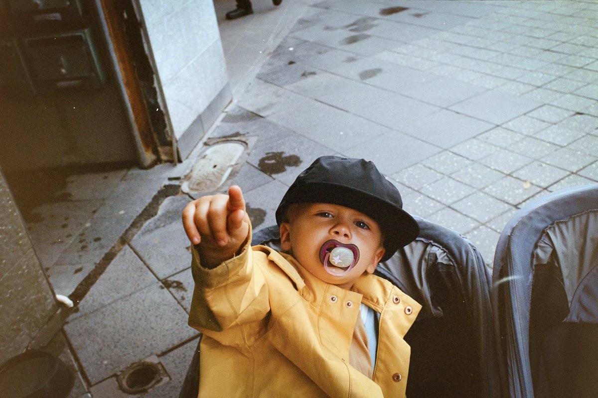 A young child sitting in a stroller, wearing a yellow jacket and black cap, with a pacifier in their mouth, pointing towards the camera.