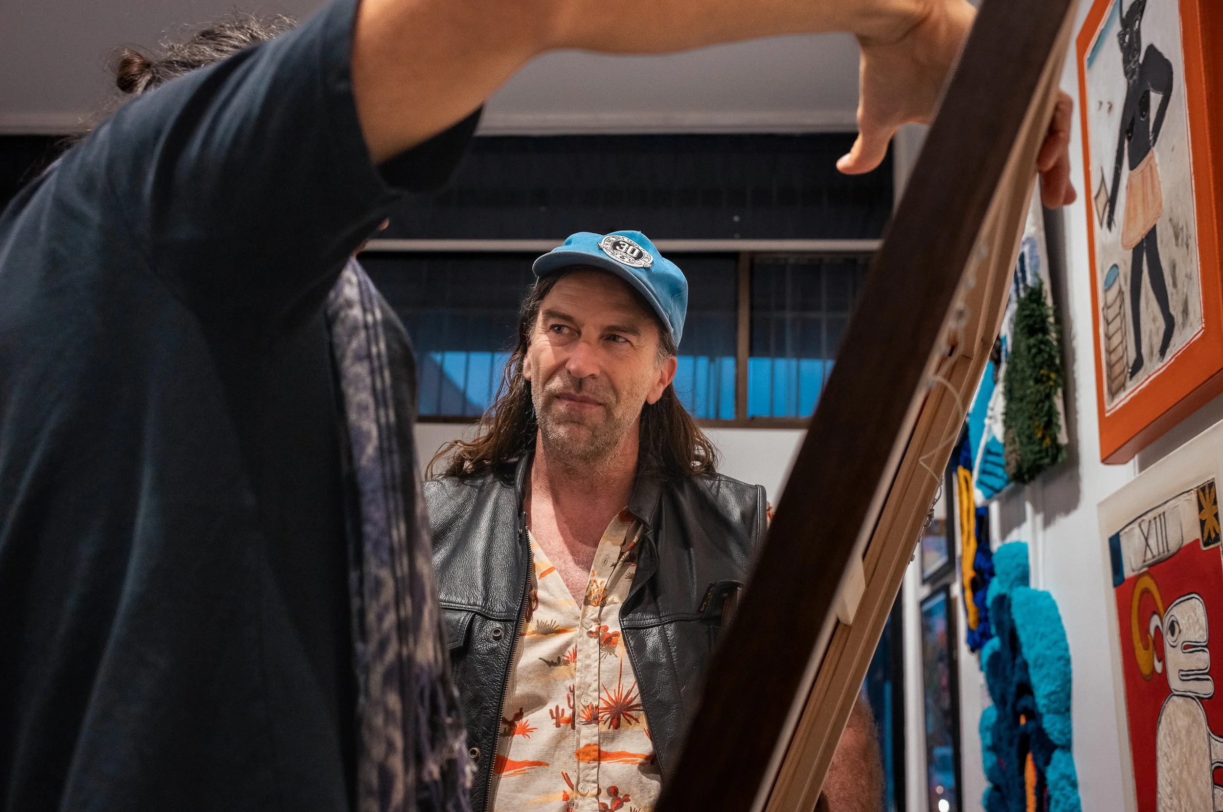 Two people looking at a painting or artwork in an art gallery or exhibit. The person on the right wears a black leather vest and a floral shirt, while the person on the left wears a cap and a jacket. The background shows colorful artwork and decorati