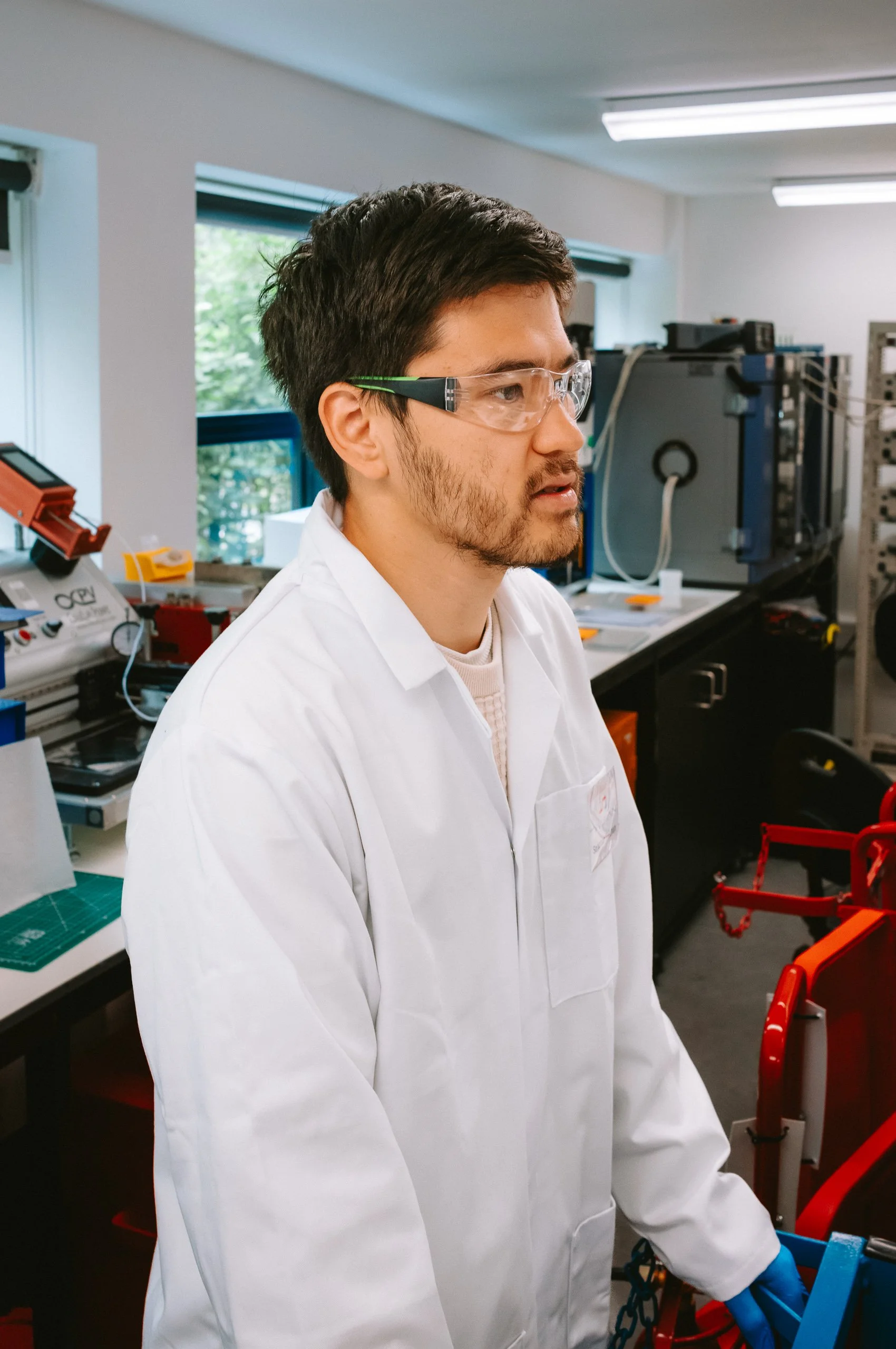 A young man wearing safety glasses and a white lab coat working in a laboratory with scientific equipment and tools.