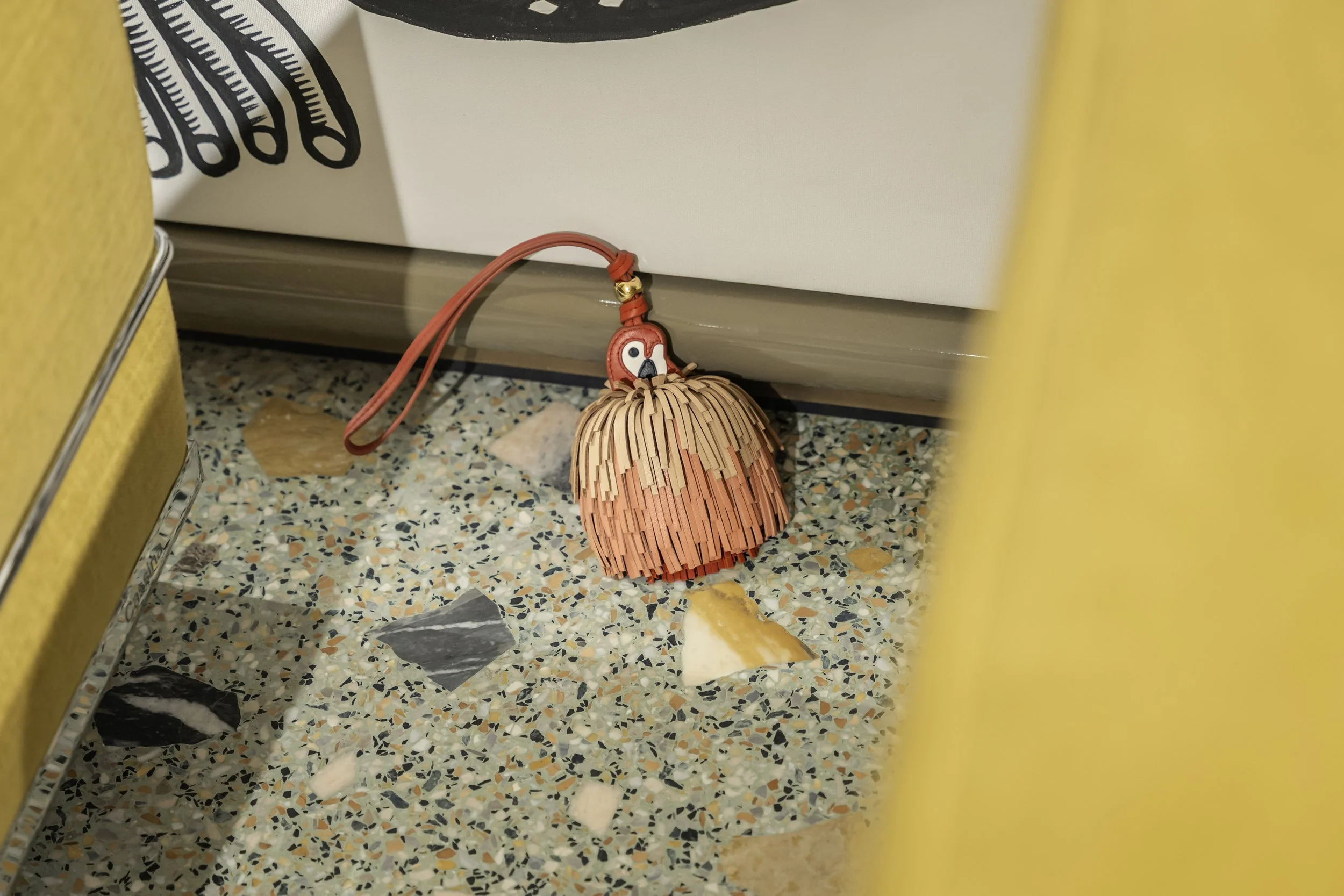 A small decorative pink purse with a straw skirt design, featuring a flamingo charm, lying on a tiled floor near a wall and some objects partially visible on the sides.