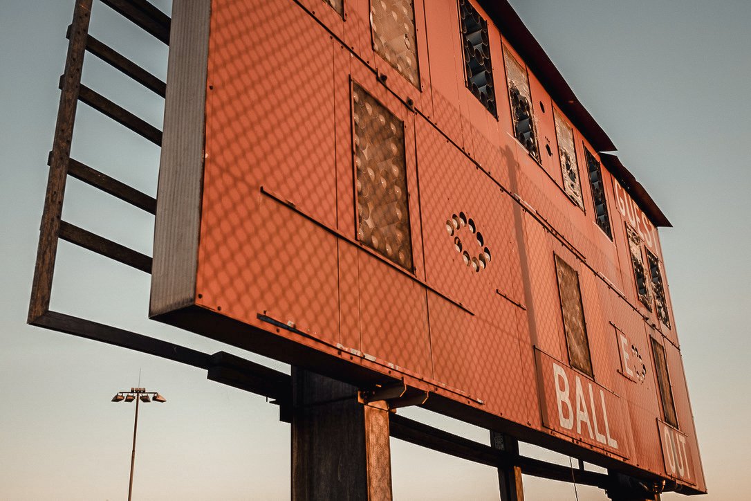 A weathered, red football scoreboard with boarded-up windows, showing the words "BALL" and part of the word "SCORES" at the top, standing on metal supports under a clear sky.