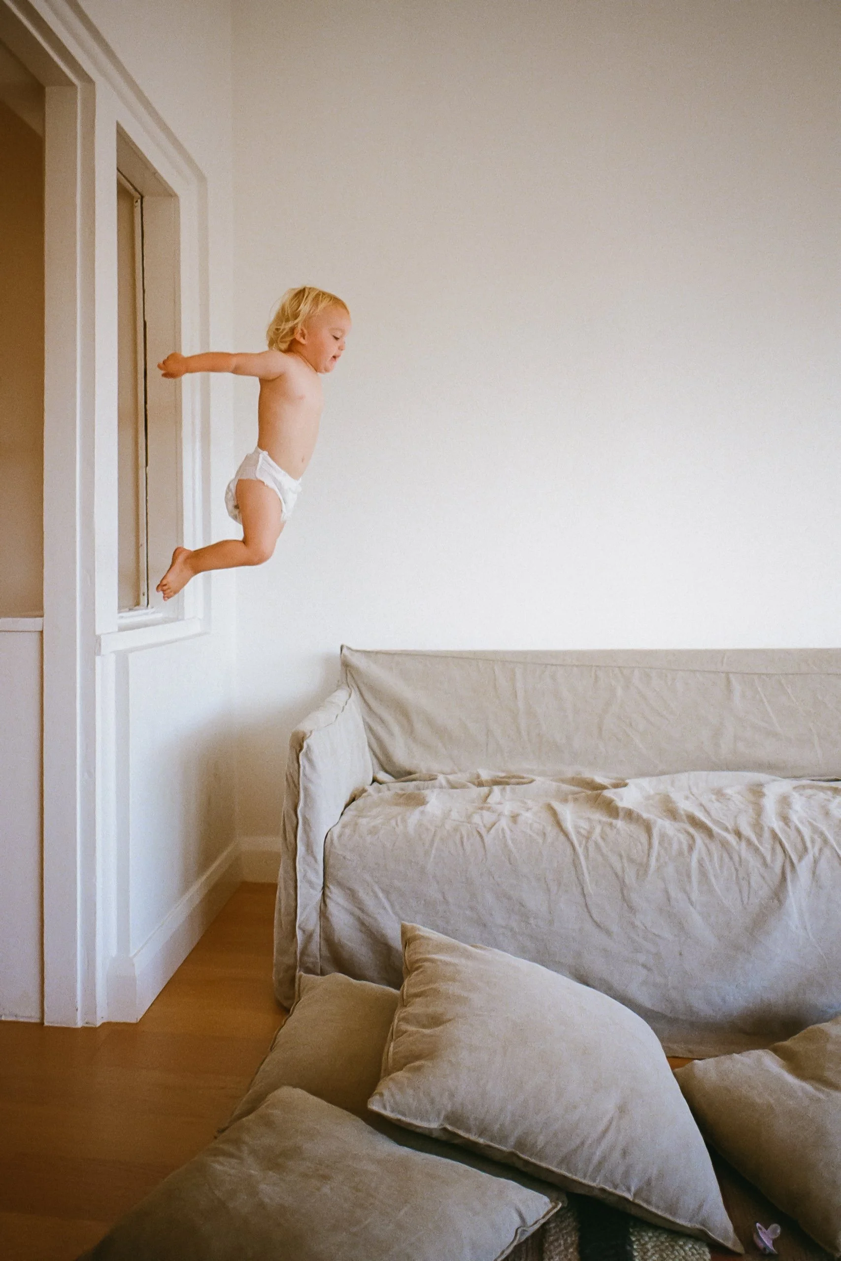 A young child jumping off a window ledge into a pile of cushions and pillows on a beige couch.