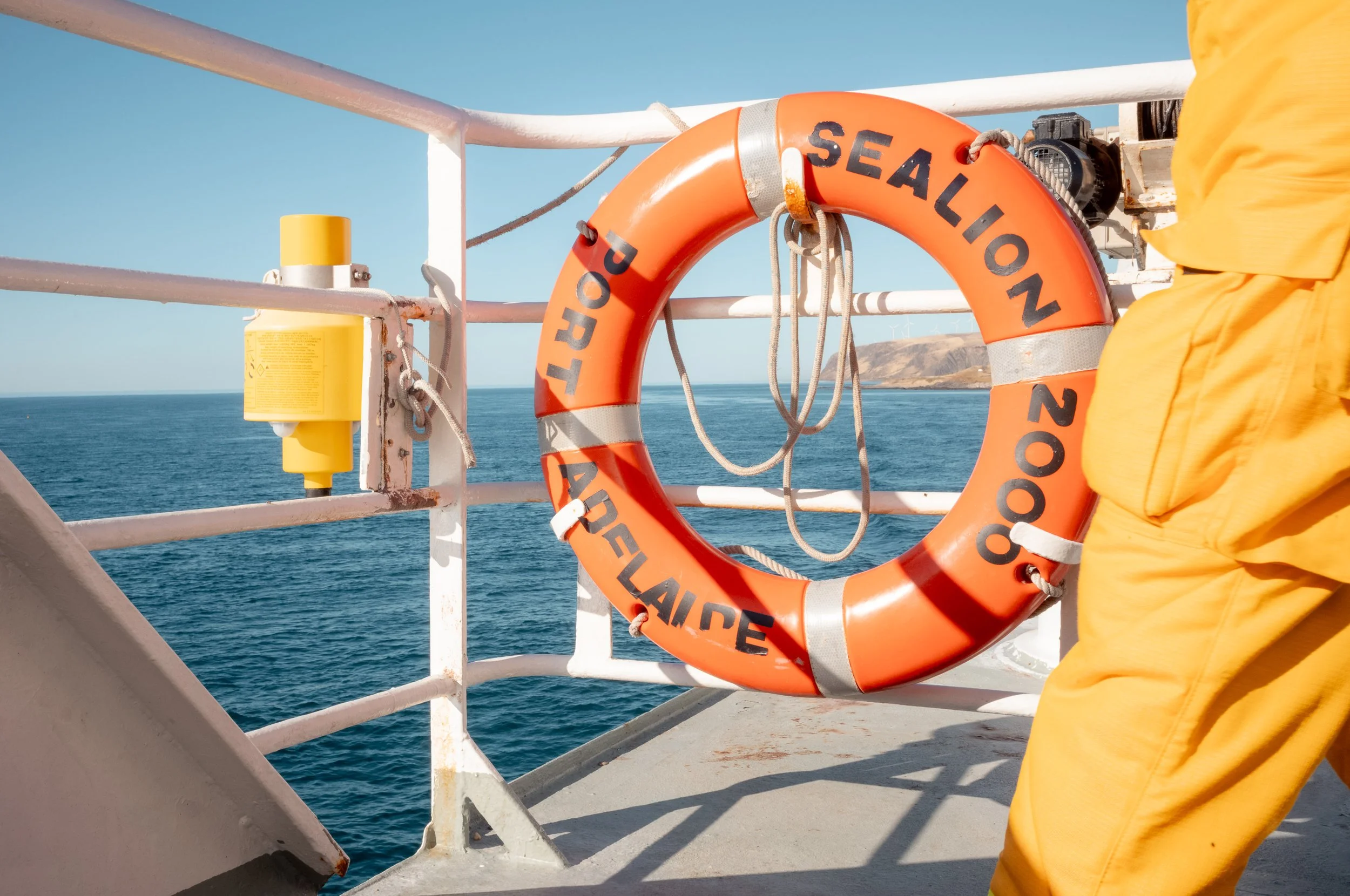 Life preserver with the words 'Port Seallon, Aukland 2009' hanging on the side of a ship overlooking the ocean, with a person in yellow clothing partially visible on the right.