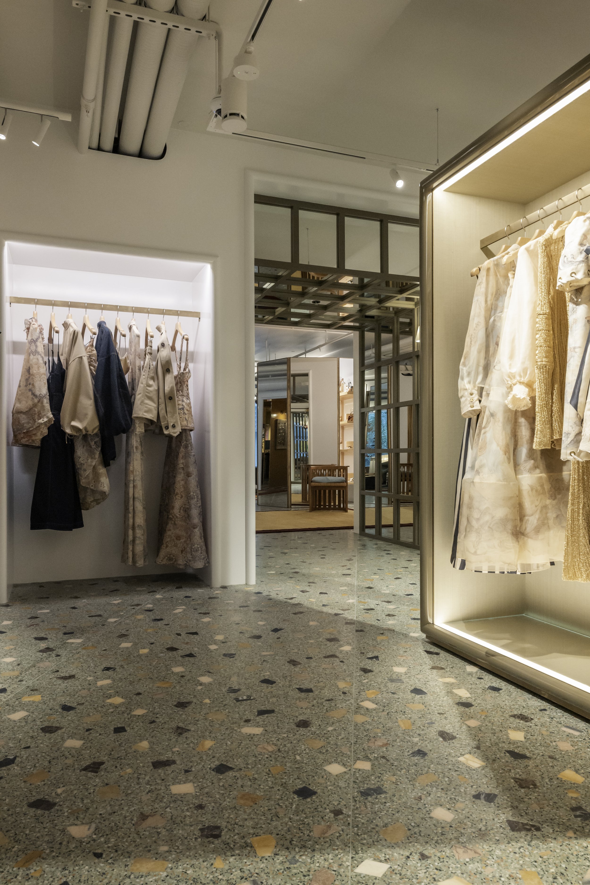 Interior of a clothing store with racks of women's clothing on either side, a terrazzo floor, and a view into another section of the store with seating and display shelves.