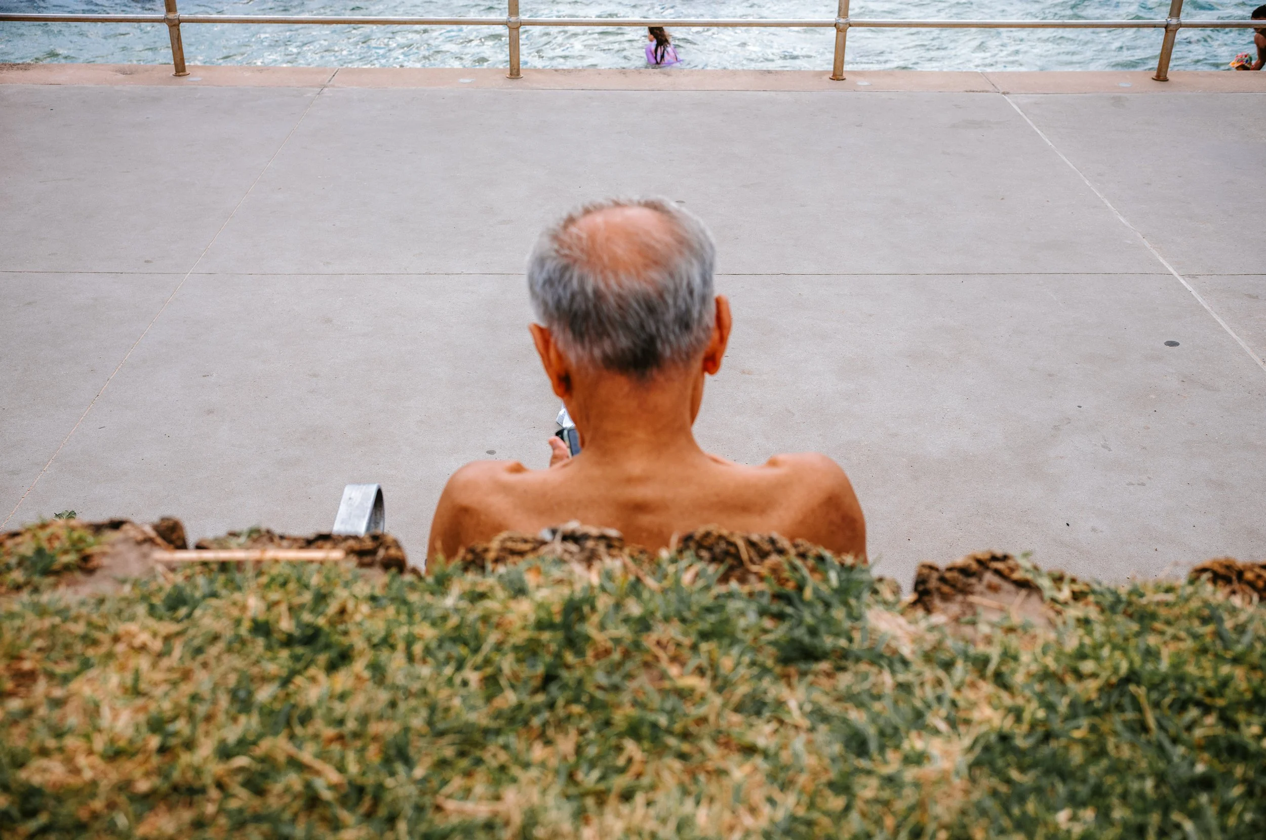 An elderly man with gray hair is sitting shirtless on a bench, facing a concrete walkway and in front of a railing overlooking a body of water. Two children are visible in the water in the background.
