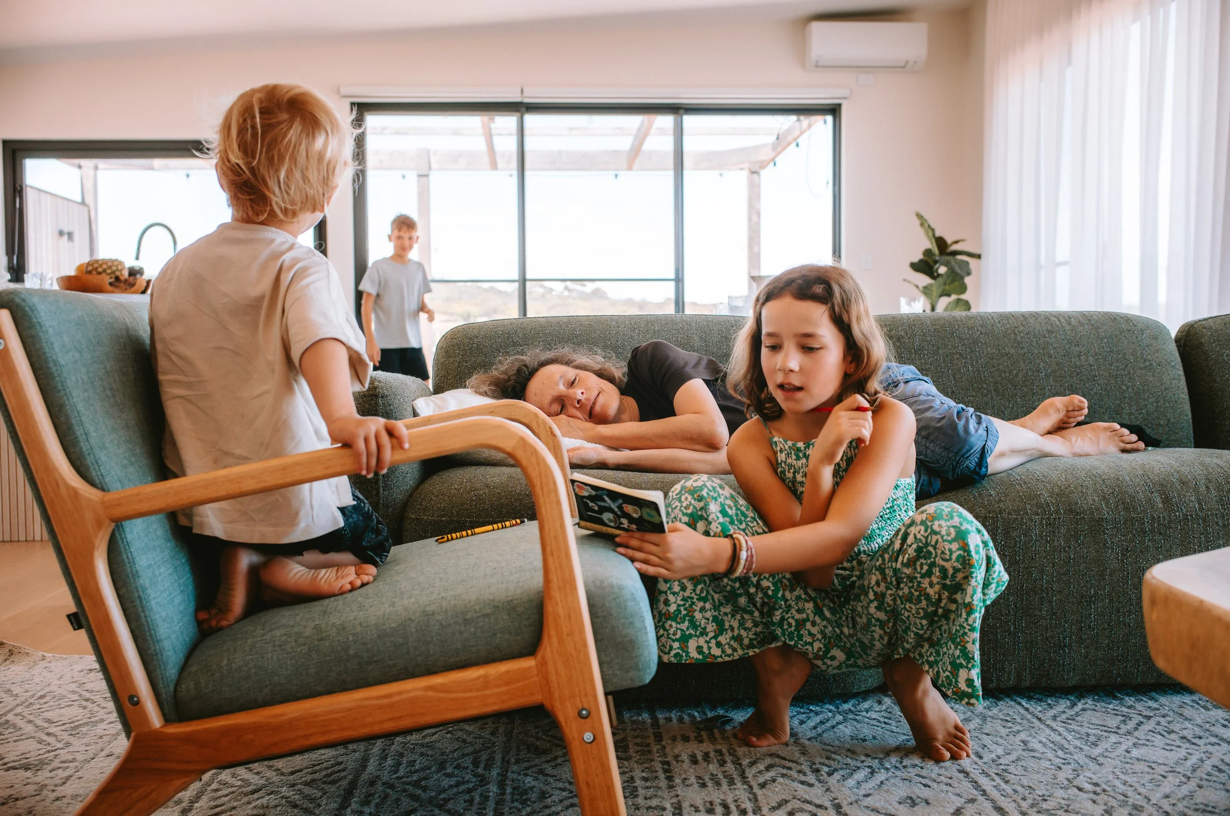 A woman lies on a couch with her head on a pillow, looking at a young girl sitting on the floor, while a boy stands in the background.
