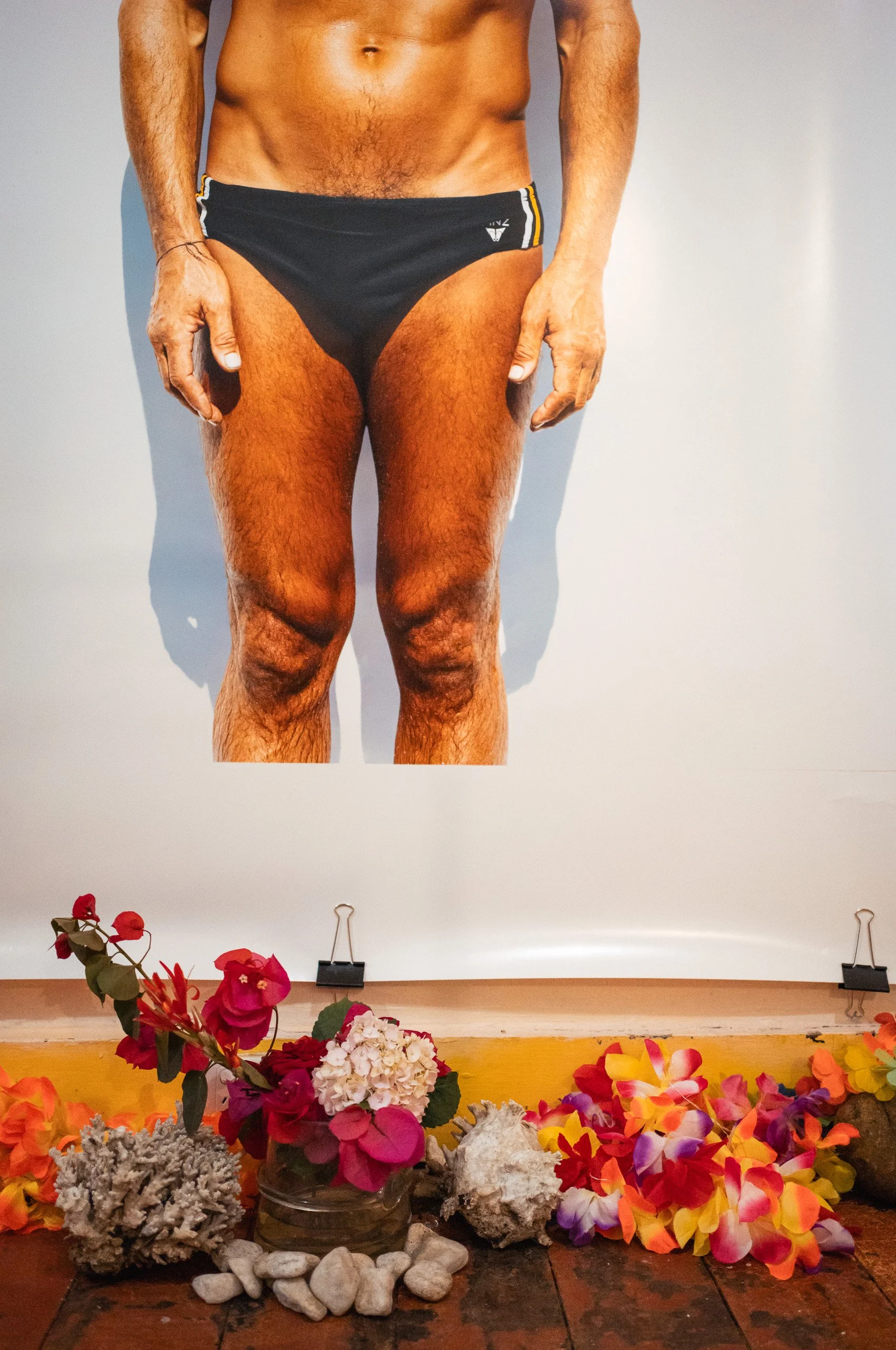 Close-up of a man wearing black swim briefs, standing against a plain light background, with flowers and rocks at his feet.