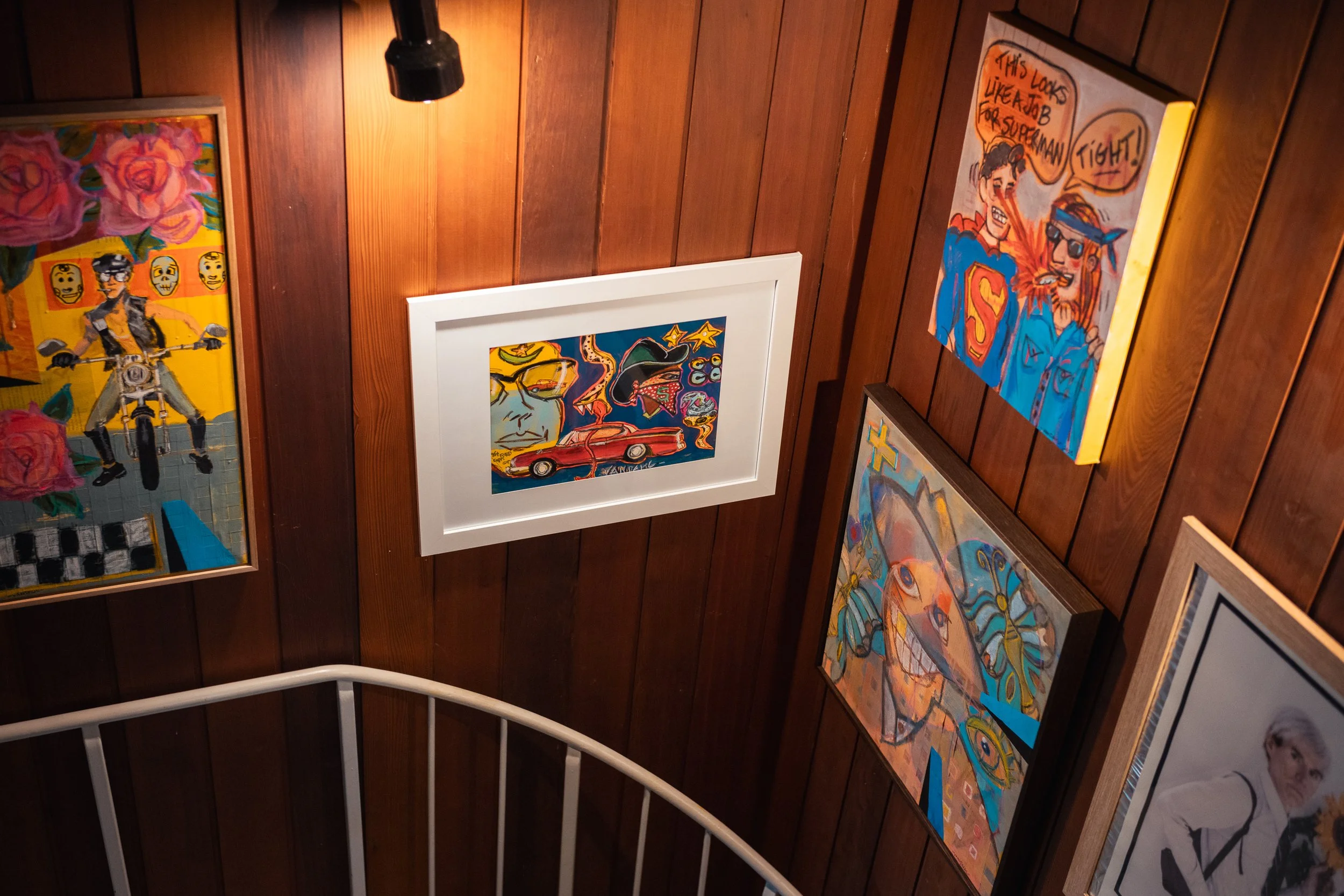 A wall covered with various colorful paintings, including portraits, abstract designs, and pop art, illuminated by a spotlight.