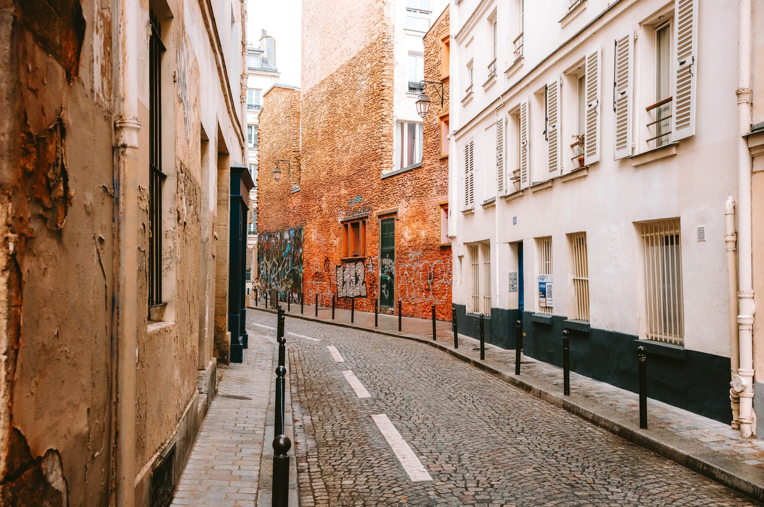 Empty cobblestone street in a European city, lined with aged buildings with graffiti, shutters, and small balconies.