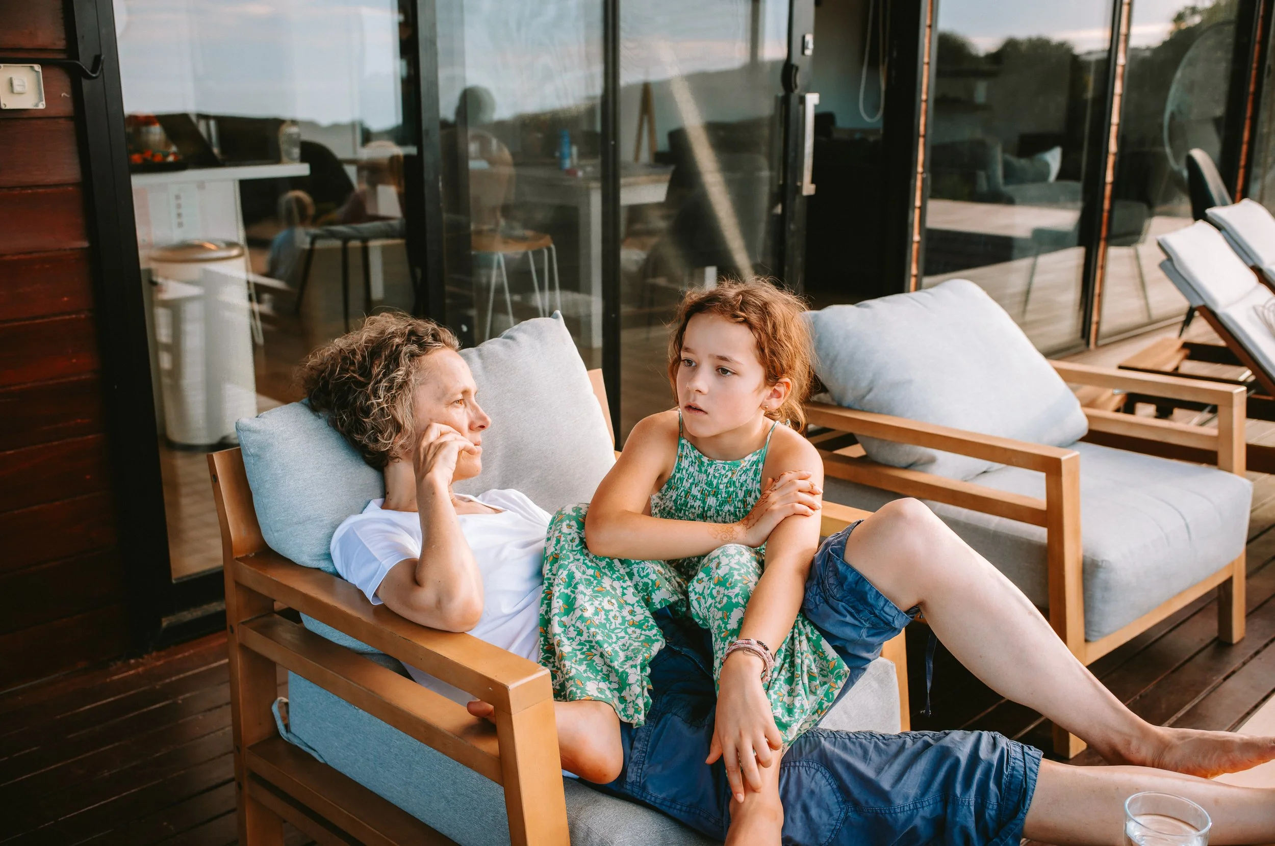 An older woman and a young girl sitting on a outdoor sofa, having a conversation. The woman is lying back, resting her head on a pillow, with her hand near her face. The girl, with curly red hair, is sitting beside her, leaning slightly forward with 