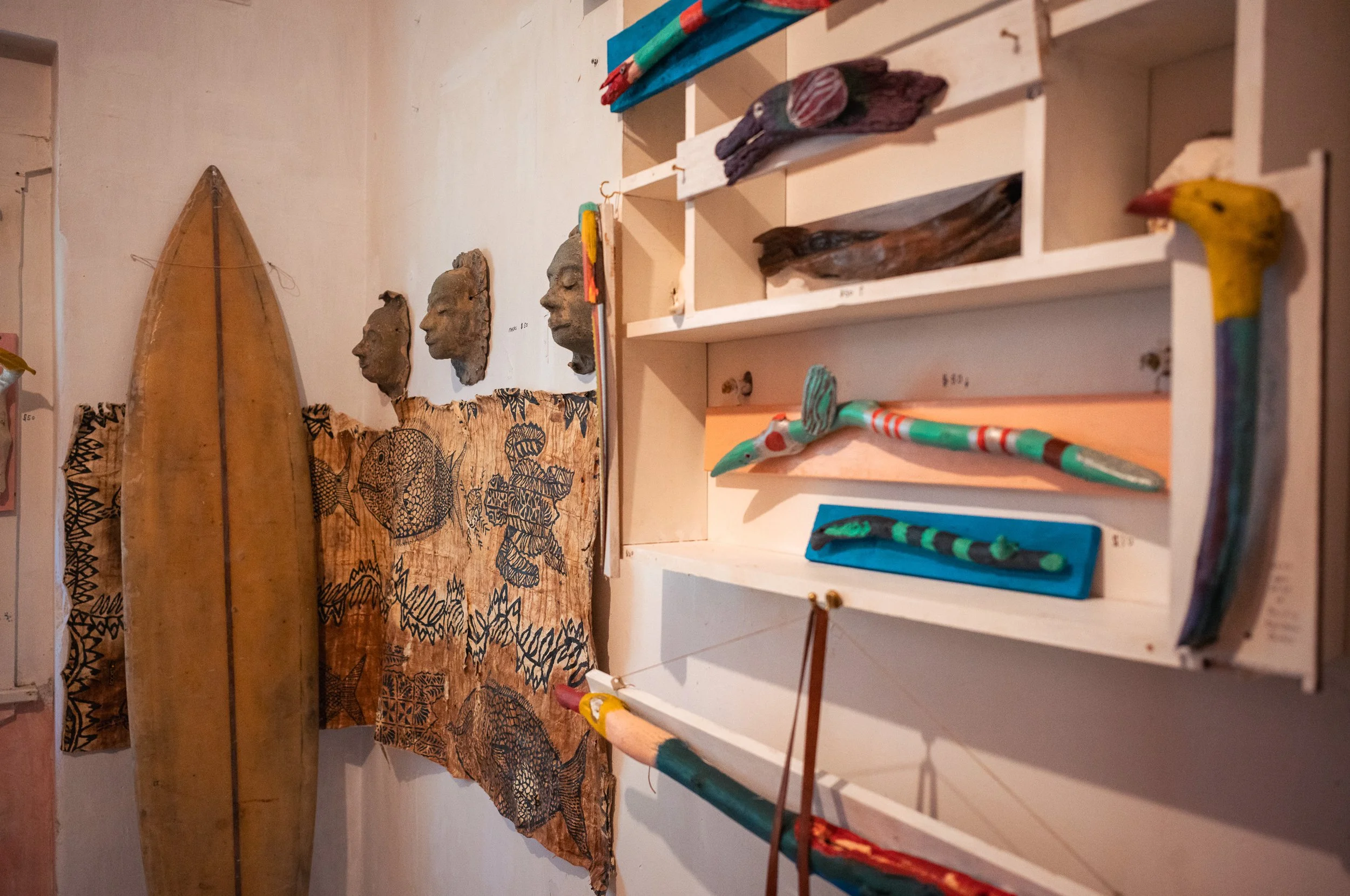 Display of African art with carved figurative heads, decorated wooden masks, colorful painted wooden animals, and a wooden boat, arranged on shelves and the wall.