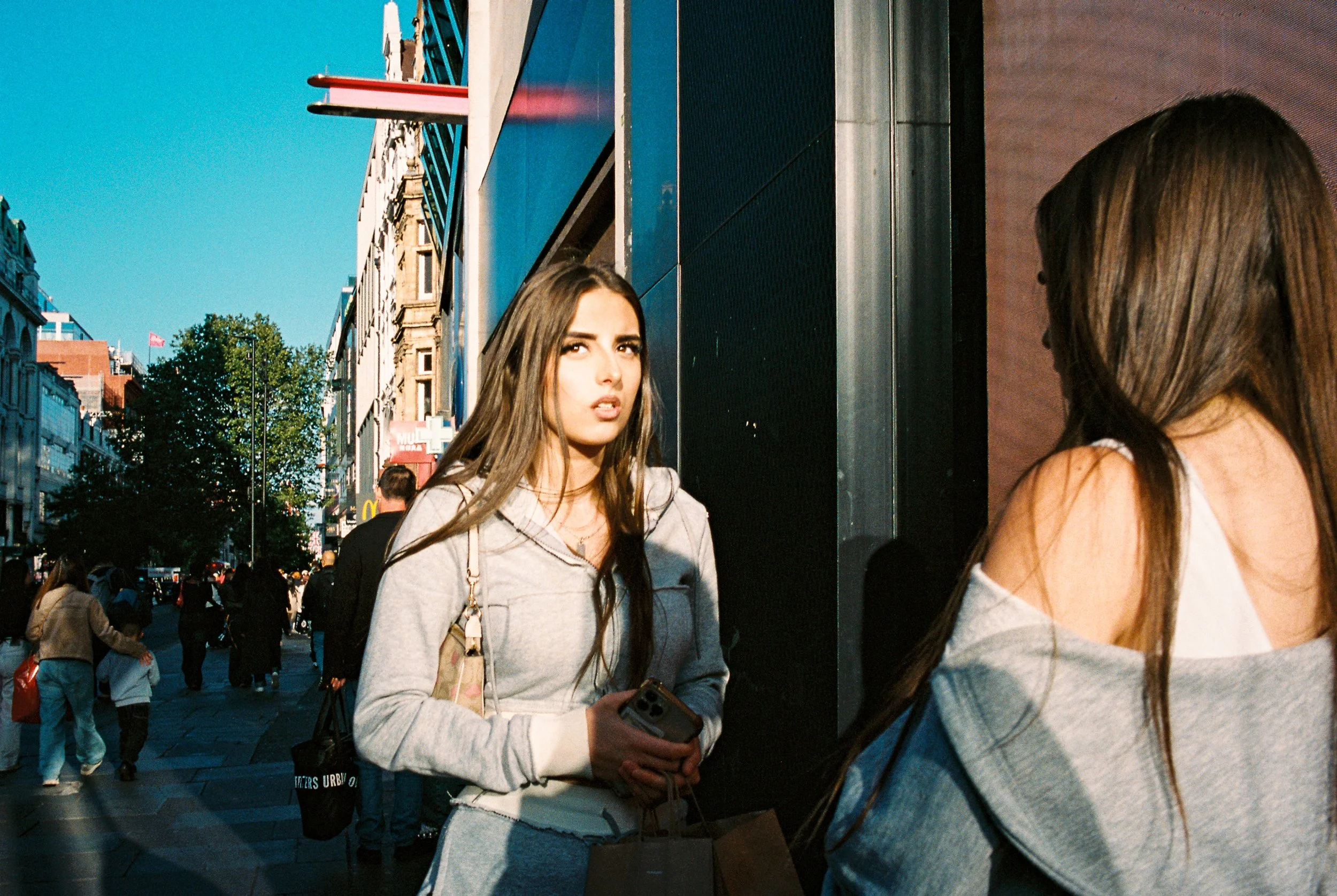 Two women standing on a busy city street, one facing the camera with a surprised expression, the other with her back turned, holding shopping bags and a phone.