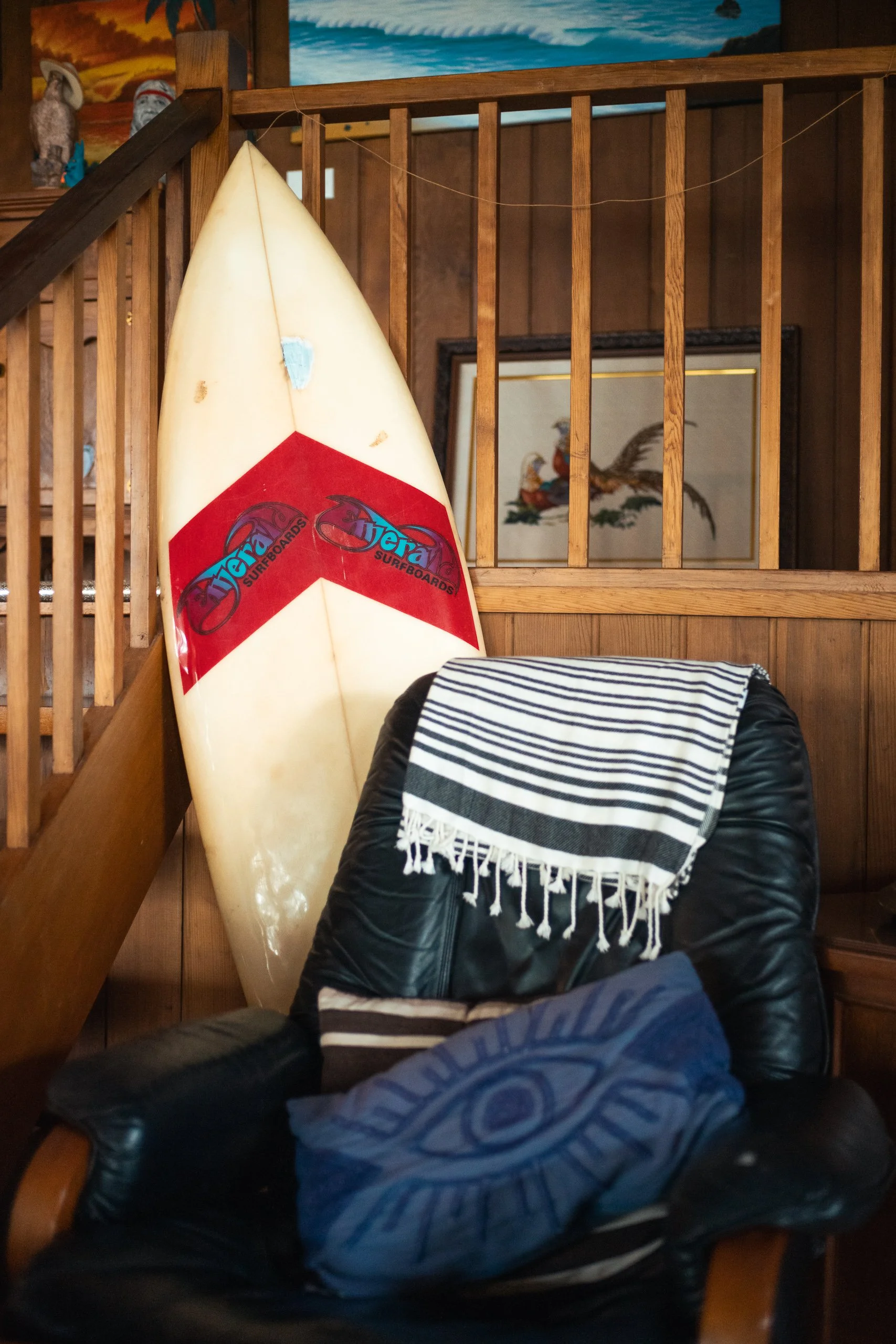A surfboard leaning against a wooden staircase railing inside a room with wooden walls and colorful wall art. A black leather armchair with a striped towel draped over the back and a blue pillow on the seat.