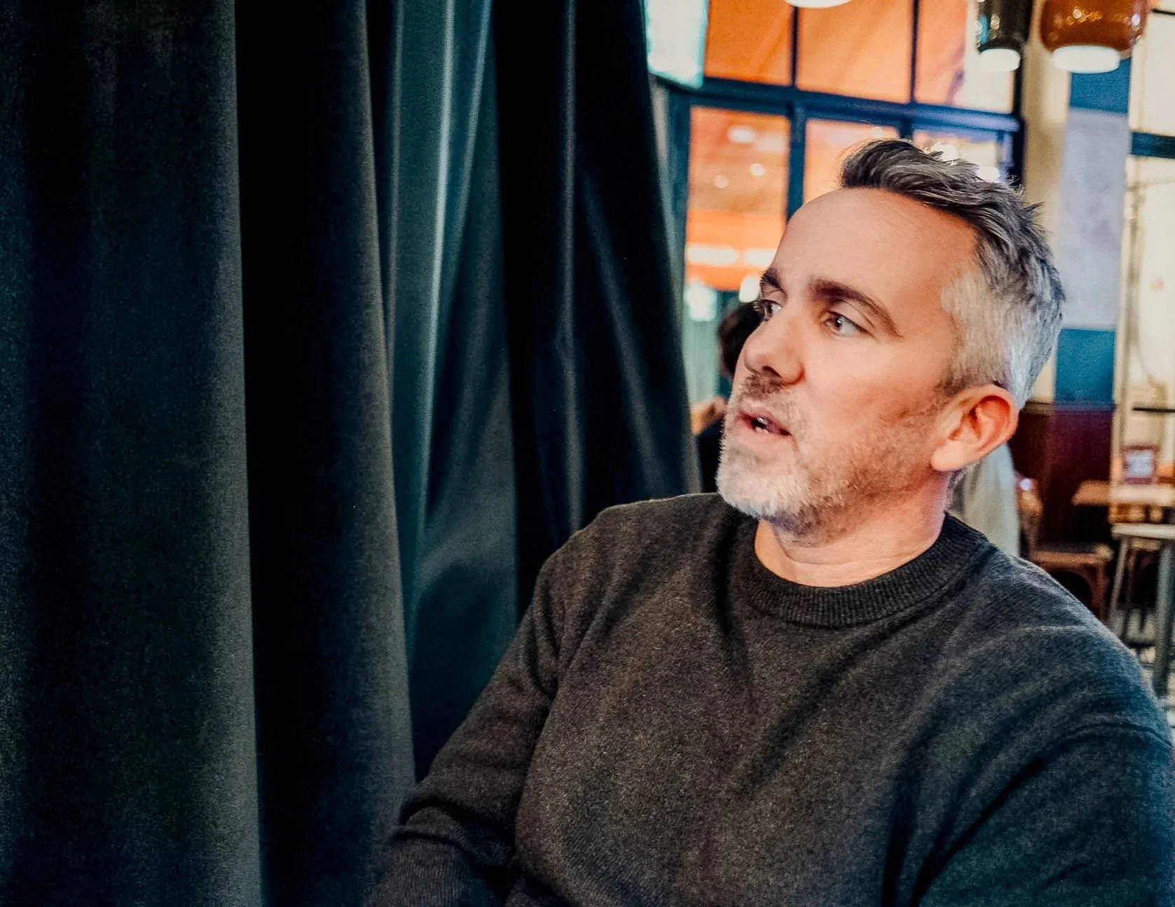 A man with gray and dark hair, wearing a dark sweater, sitting indoors near black curtains in a restaurant or cafe. He appears to be speaking or listening, with a focused expression.