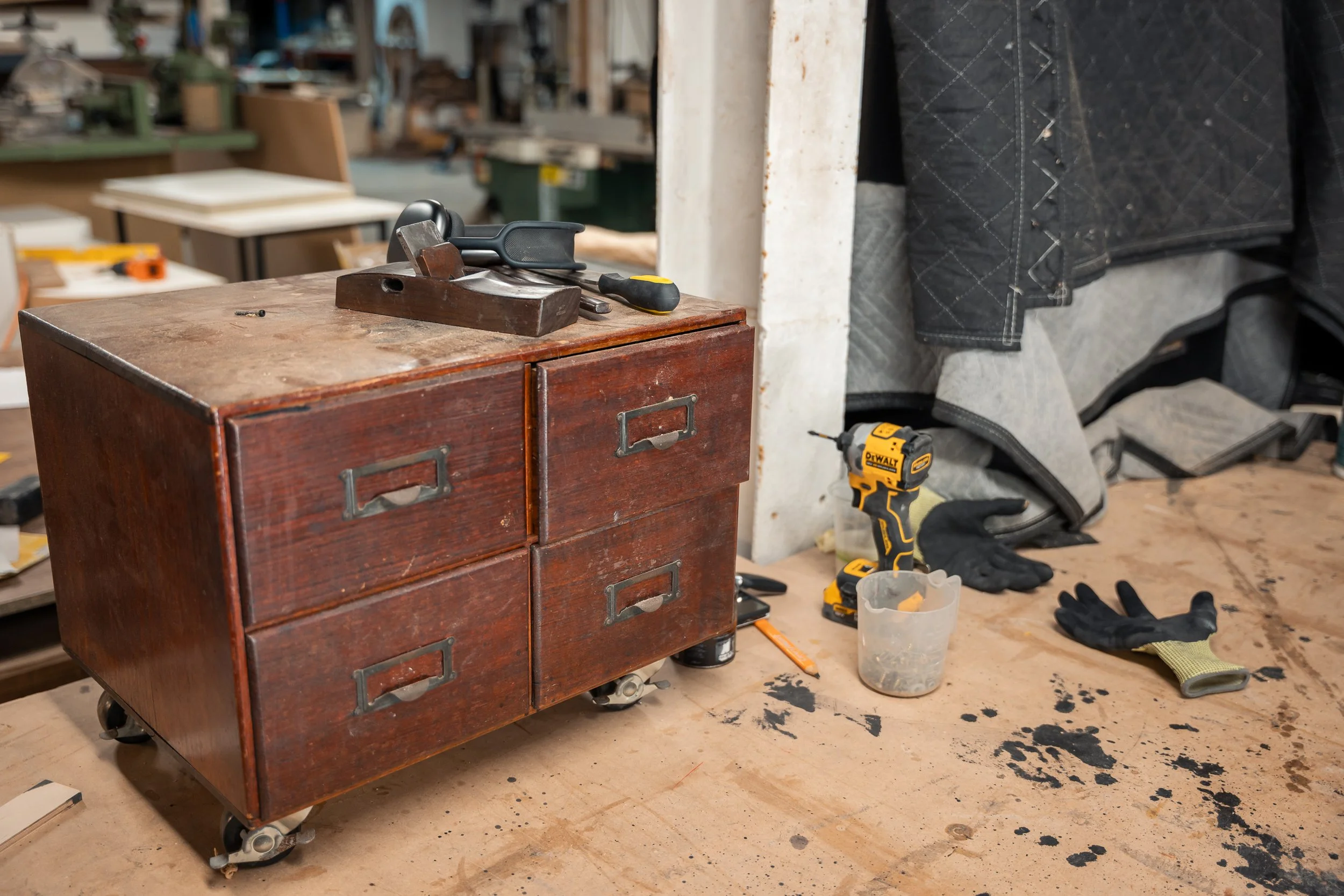 A wood workbench cluttered with black stains, a small wooden filing cabinet on wheels, a cordless drill, a screwdriver, gloves, and a small plastic container in a woodworking workshop.