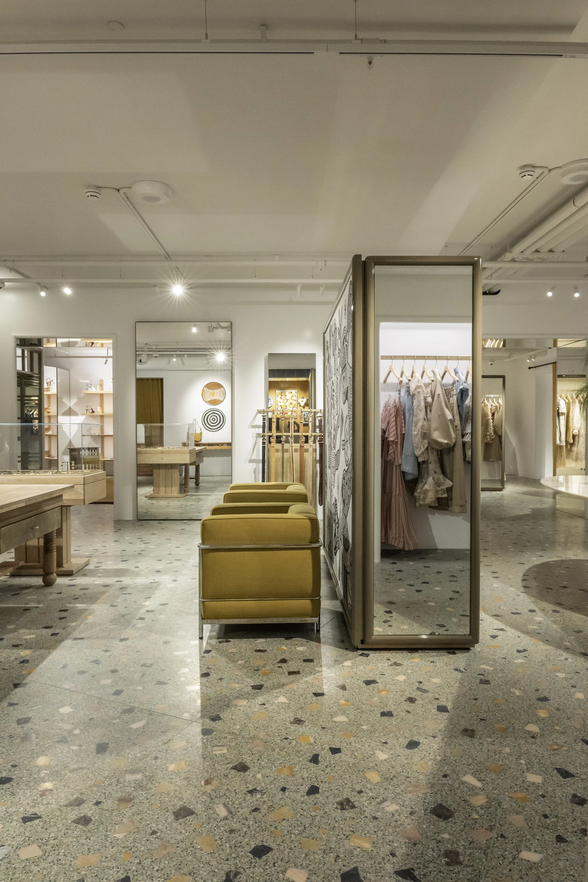 Interior of a retail clothing store displaying chairs, clothing racks, and shelving with fashion items, under bright lighting with a terrazzo floor.