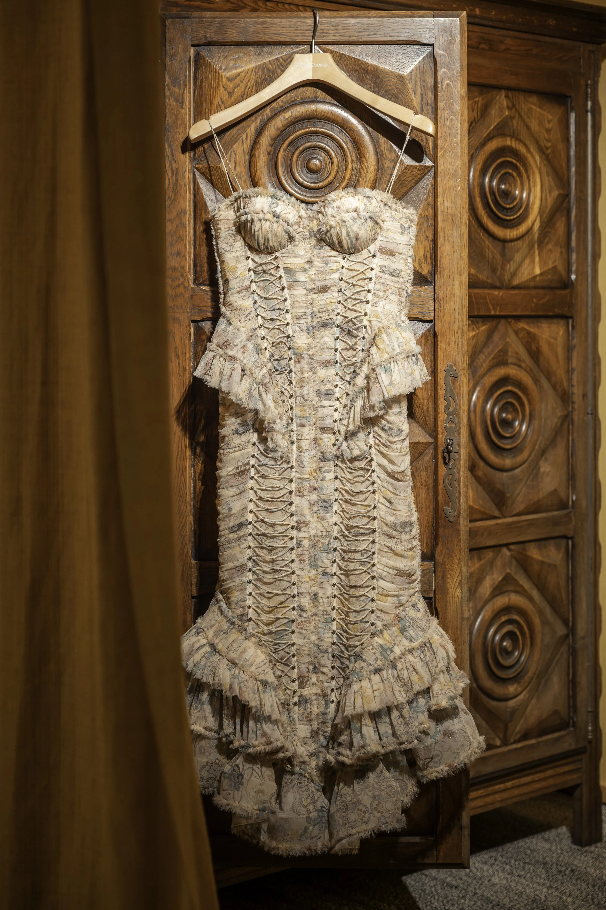 A vintage dress with intricate lace and ribbon details hanging on a wooden hanger on an ornately carved wooden wardrobe.