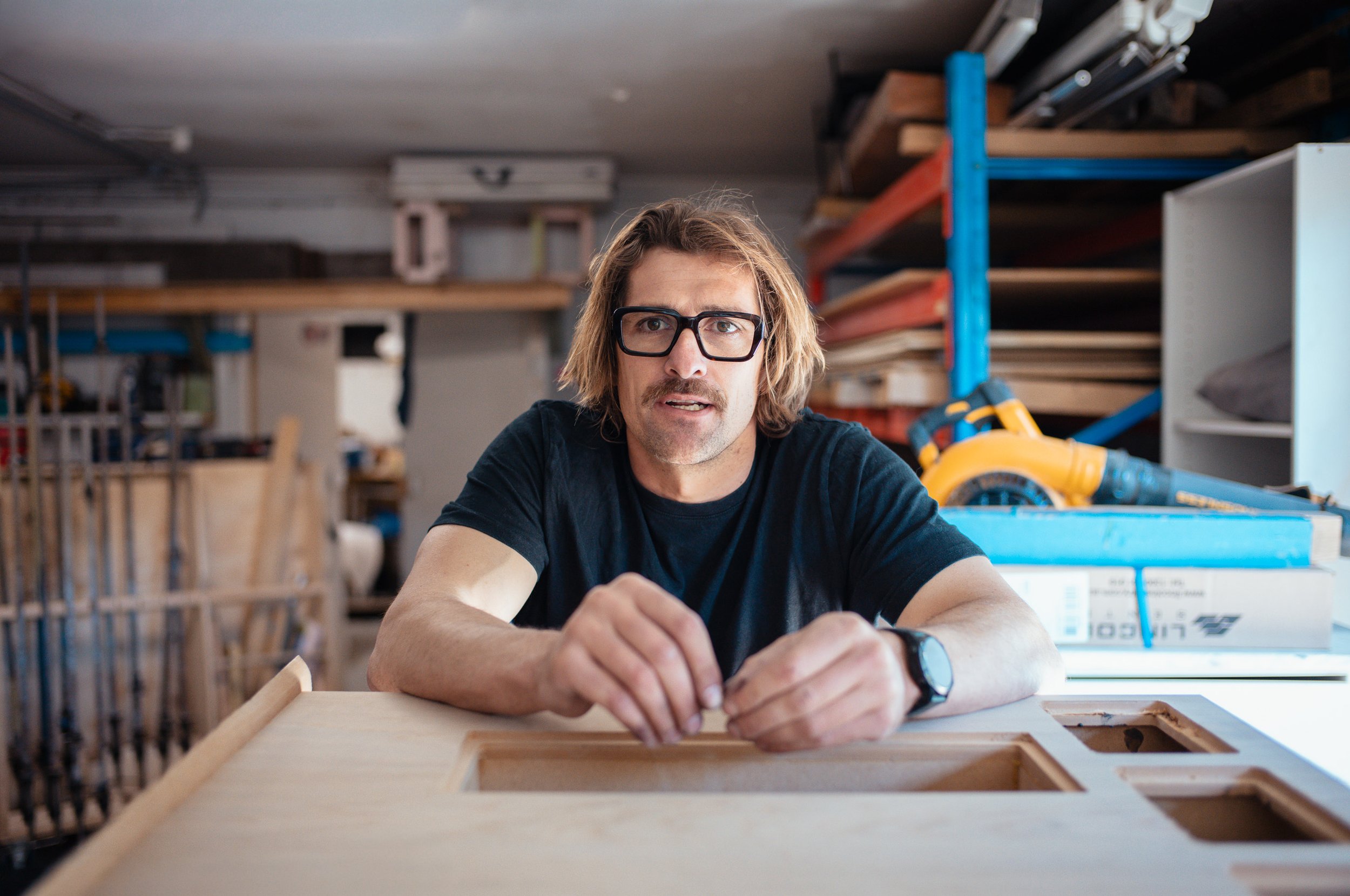 Man with glasses and a watch working on a woodworking project in a workshop filled with tools and wood pieces.