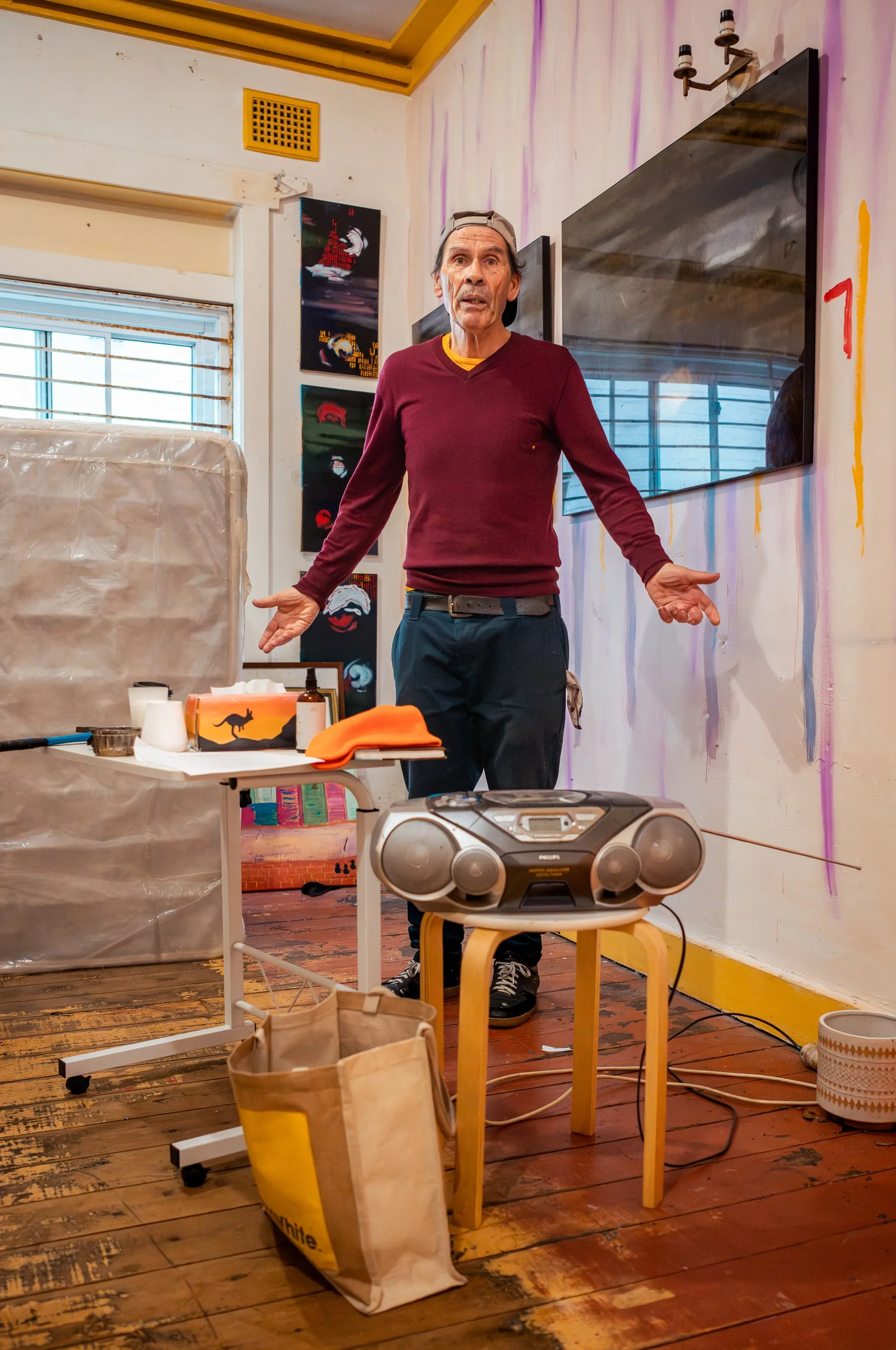 A man wearing a maroon sweater and backwards baseball cap stands in an art gallery or studio, gesturing with his hands. There is a small table with art supplies and a boombox, a large mirror on the wall, and colorful abstract paintings behind him.