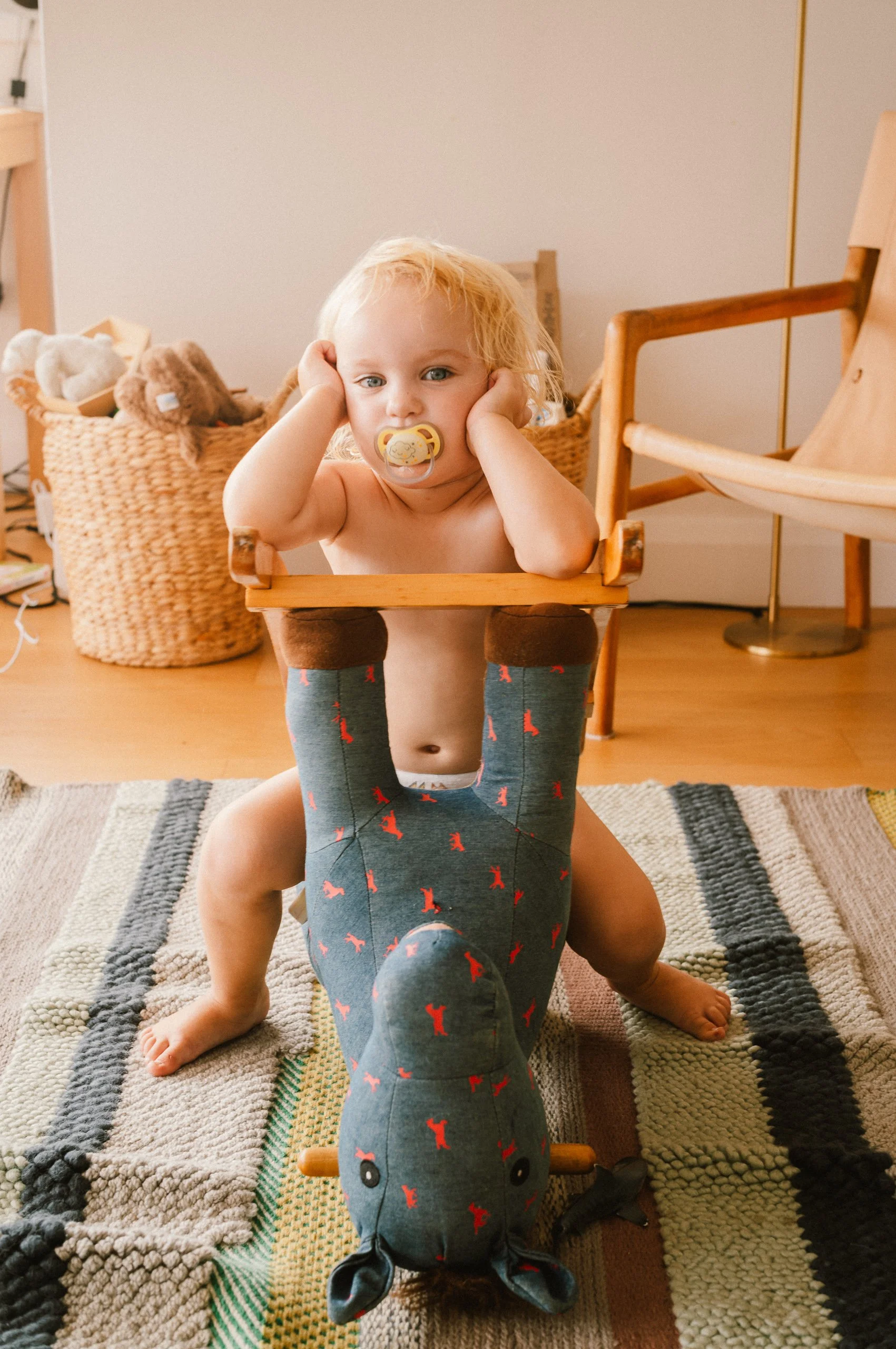 A young child with blonde hair and a pacifier in their mouth, sitting on a plush toy horse, with their arms resting on a wooden frame while looking at the camera. The room has a basket of stuffed animals in the background and a colorful striped rug o