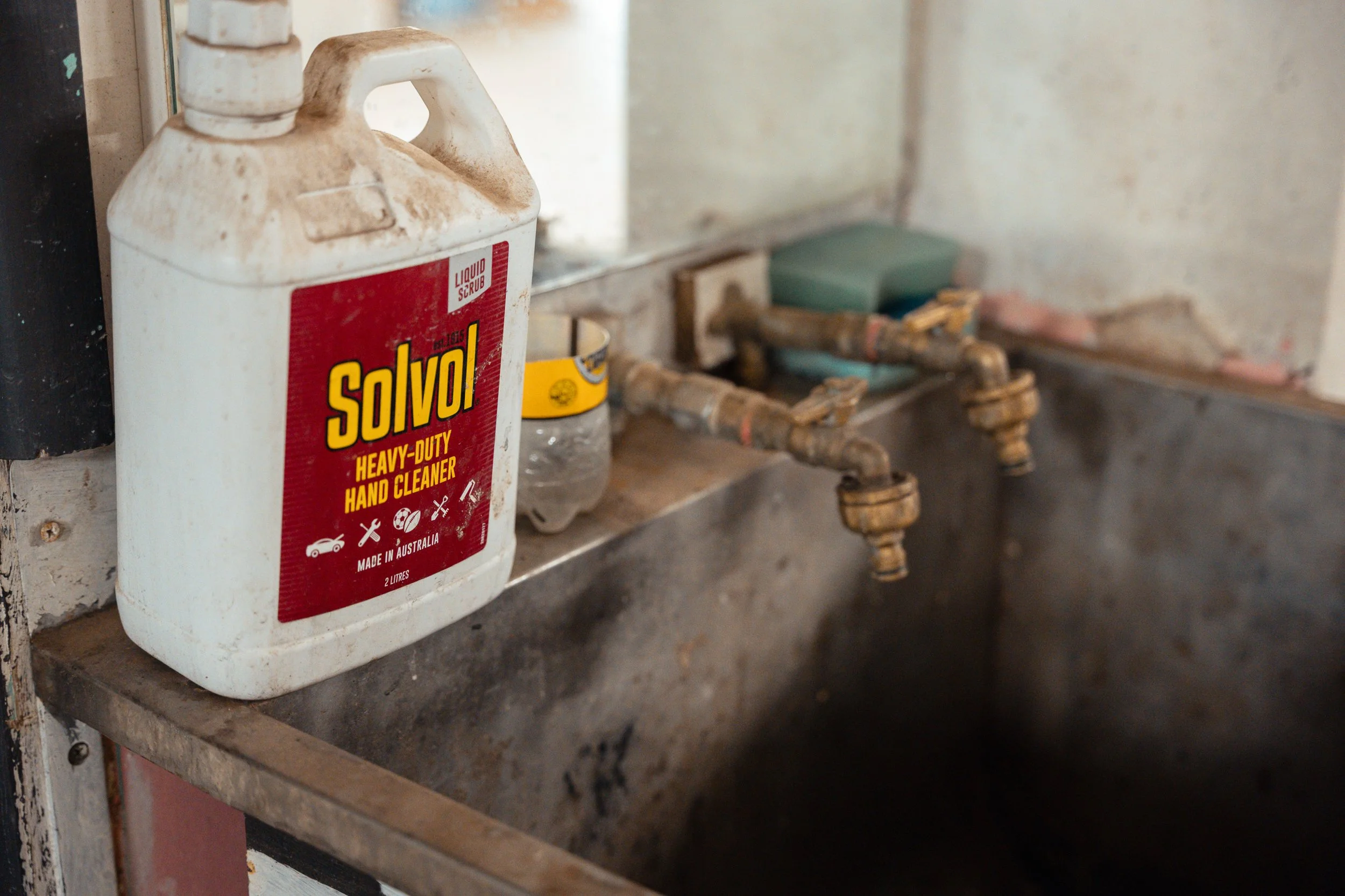 A white bottle of Solvol heavy-duty hand cleaner placed on the edge of a rusty metal sink with old, corroded plumbing fixtures in a worn, dirty industrial or workshop setting.