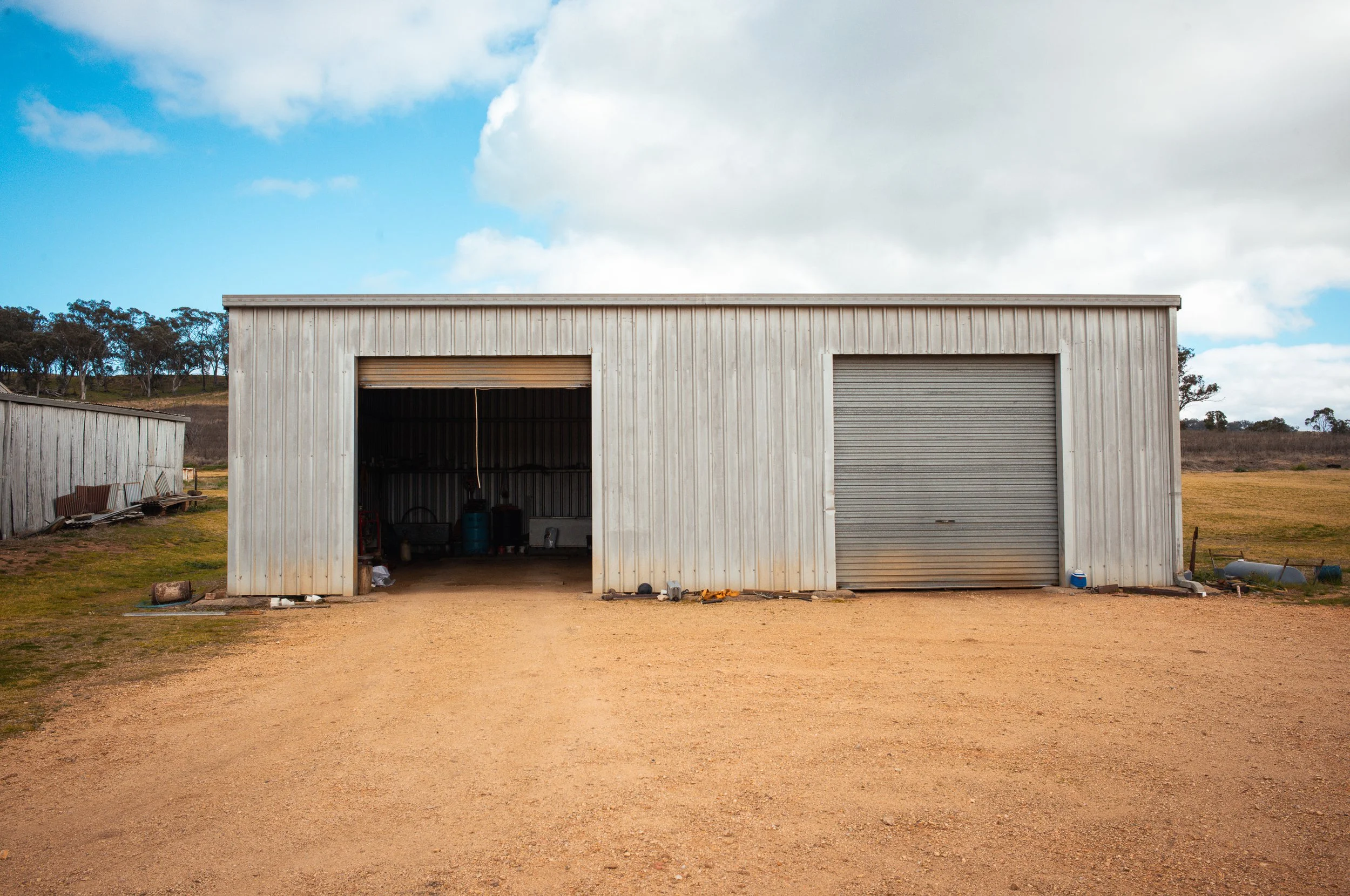Metal shed with two large roll-up doors, one partially open showing equipment inside, situated in a rural area with a dirt driveway, grassy fields, and trees in the background under a partly cloudy sky.