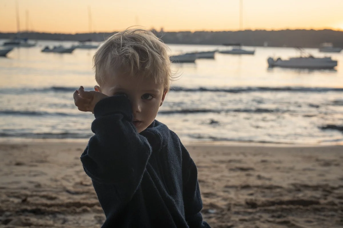 A young boy with blonde hair and a dark jacket standing on a beach at sunset, with boats on the water in the background.