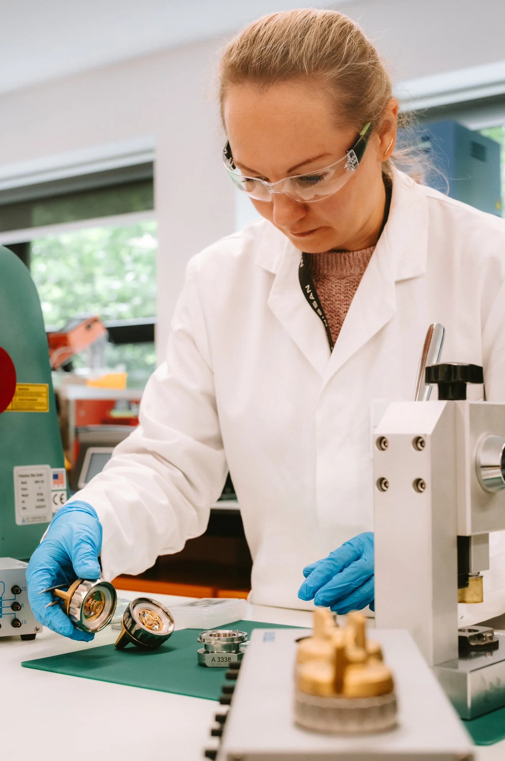 A scientist in safety glasses and gloves examines a mechanical part in a laboratory.