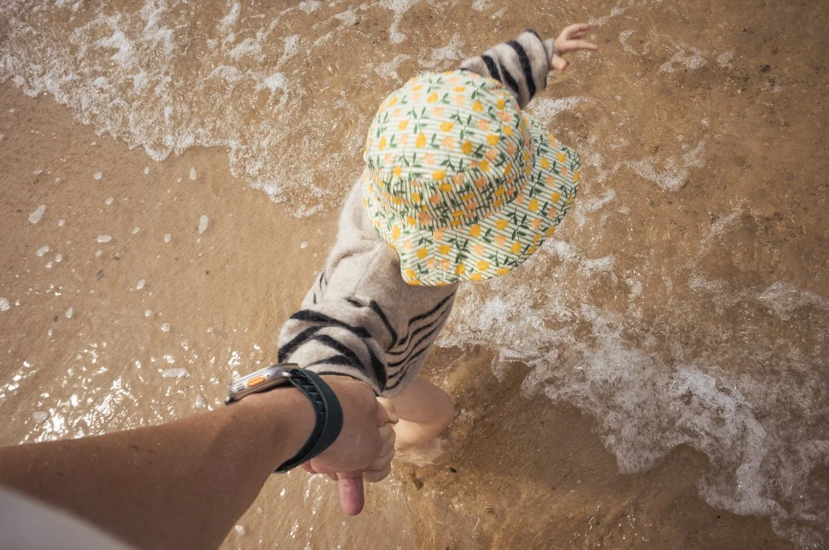 An adult holding a child's hand walking in the shallow surf at the beach, with the child's head turned away wearing a patterned hat.