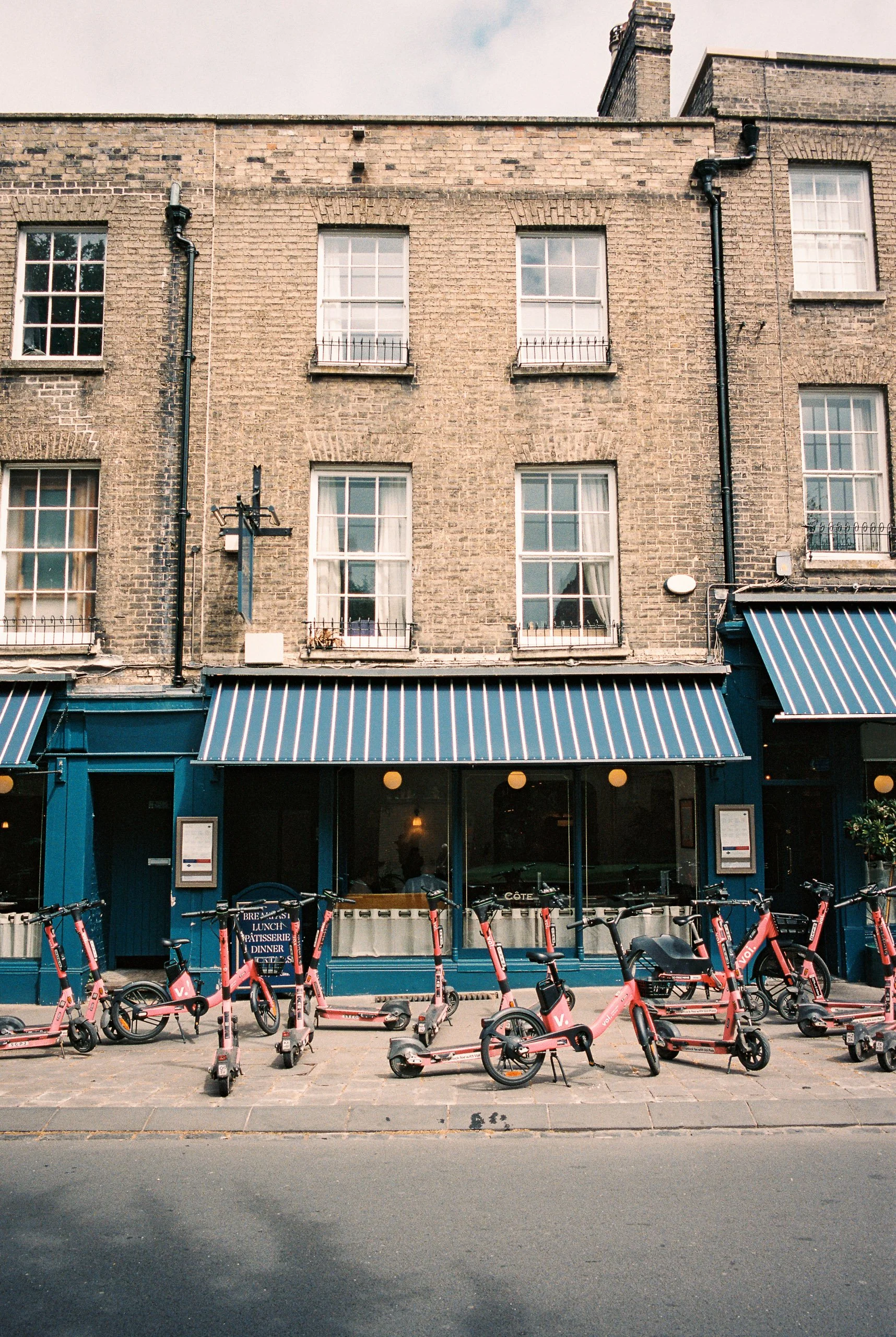A row of pink and black scooters parked outside a storefront with blue walls and striped blue and white awnings, above which are brick building windows.