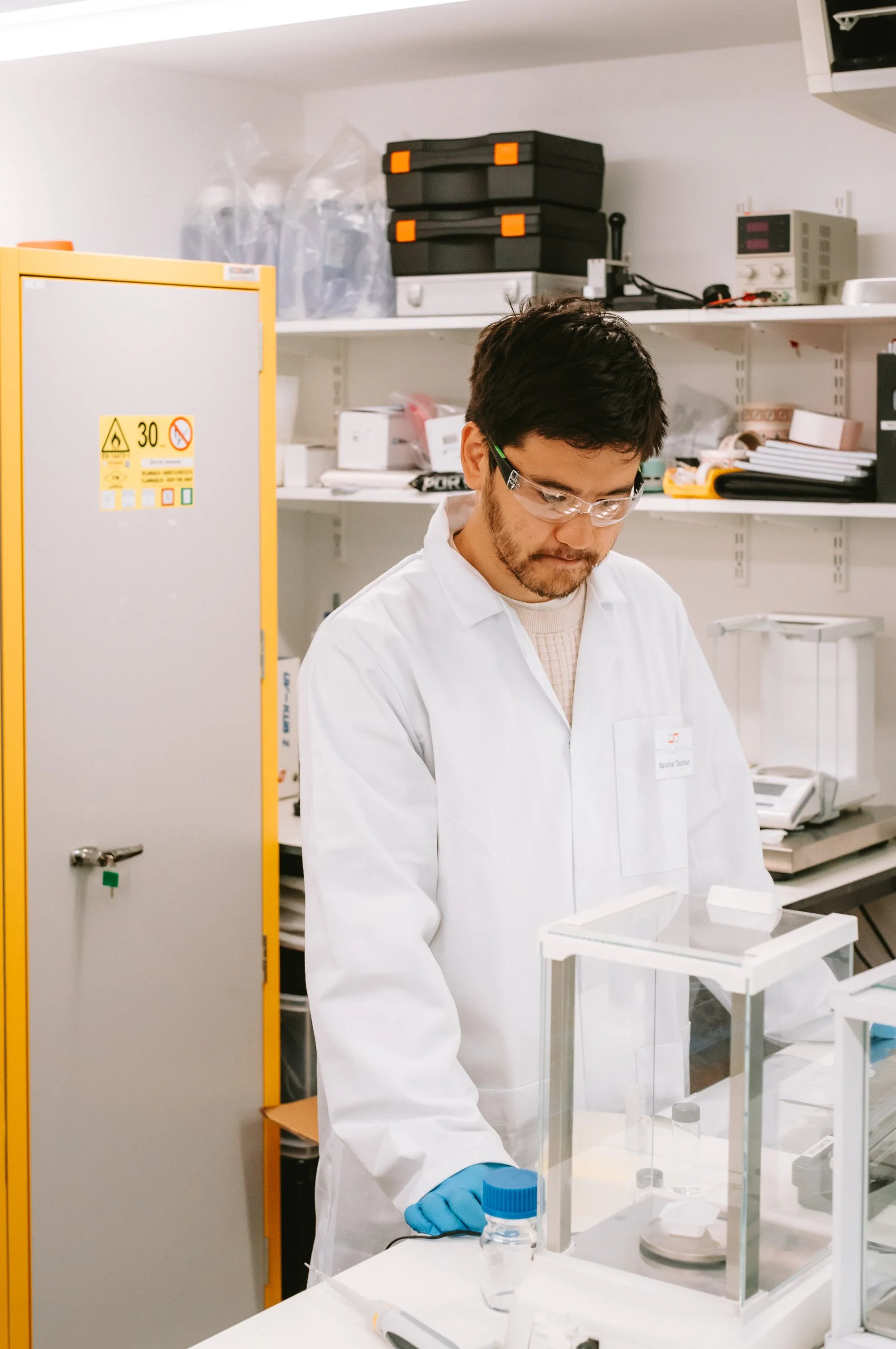 A scientist in a lab coat and safety goggles working in a laboratory, handling a small container with a blue cap.
