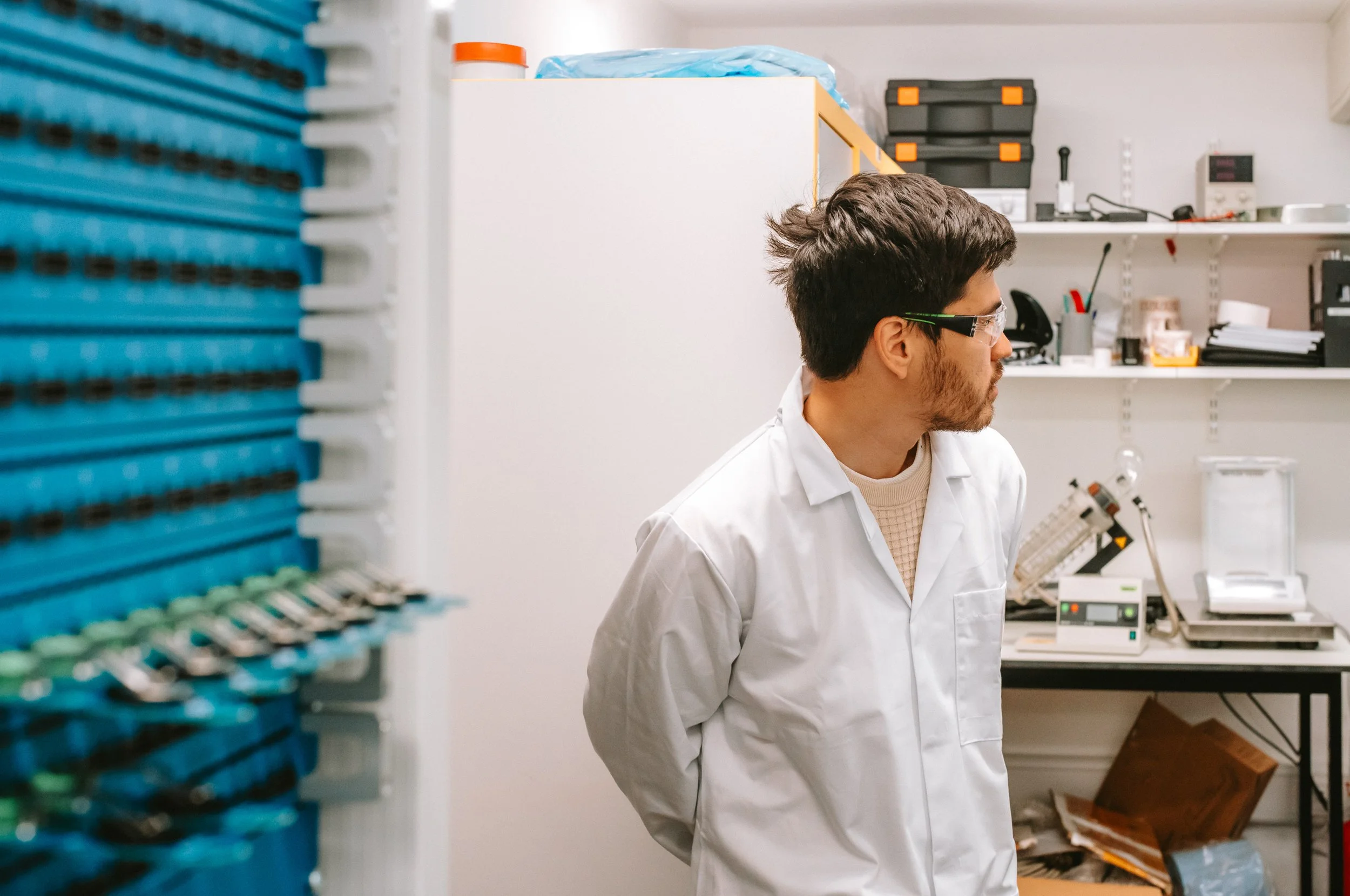 A male scientist in safety glasses and a white lab coat standing in a laboratory with shelves and scientific equipment in the background.