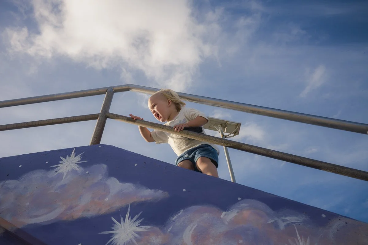 A young child with blonde hair, wearing a white shirt and denim shorts, stands on a slide at a playground, yelling or crying. The slide has a painted galaxy scene with stars and planets. The sky is blue with some clouds.