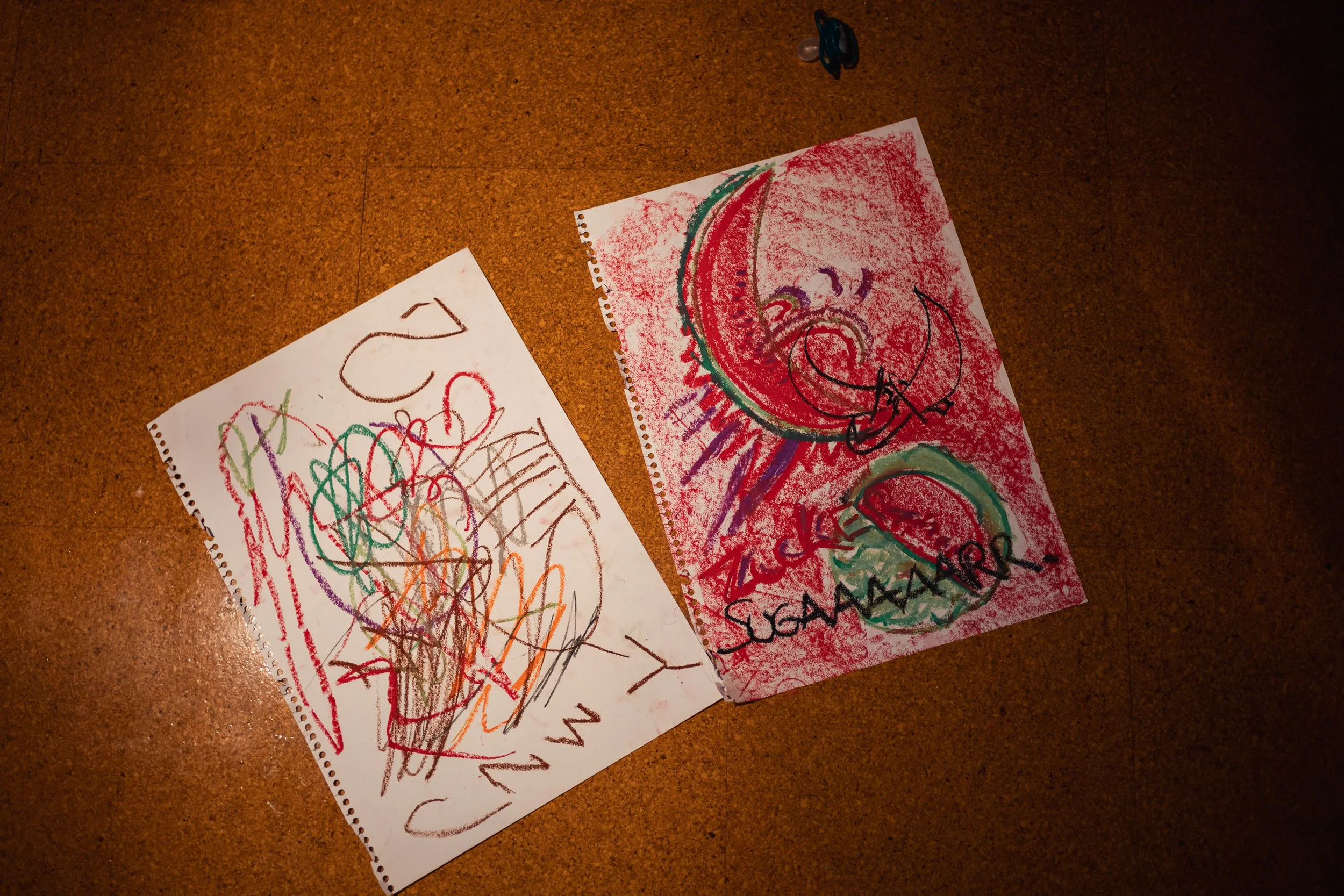 Two pieces of children's drawings on a brown floor. The left drawing has colorful scribbles, and the right one has a red background with a drawing of a face, a crescent moon, and the word "SQUASH" written on it.