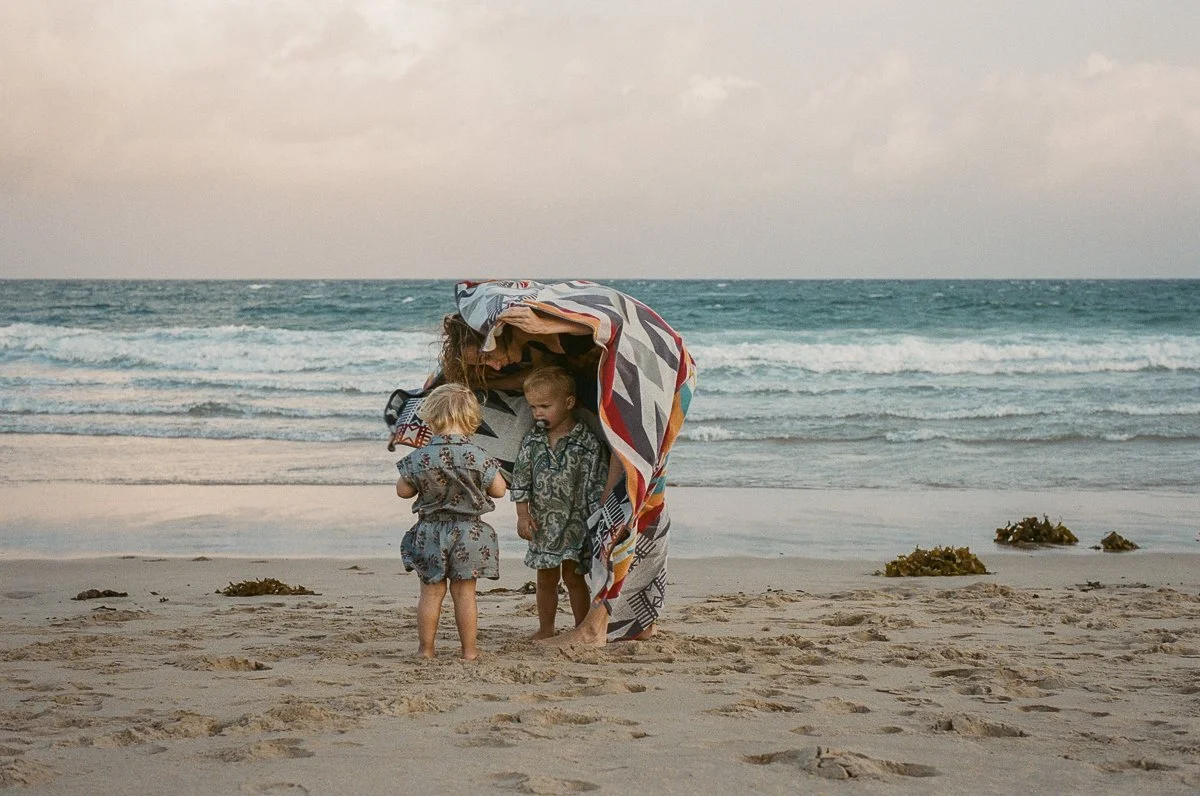 A woman and three children standing under a colorful towel on a sandy beach with the ocean in the background.
