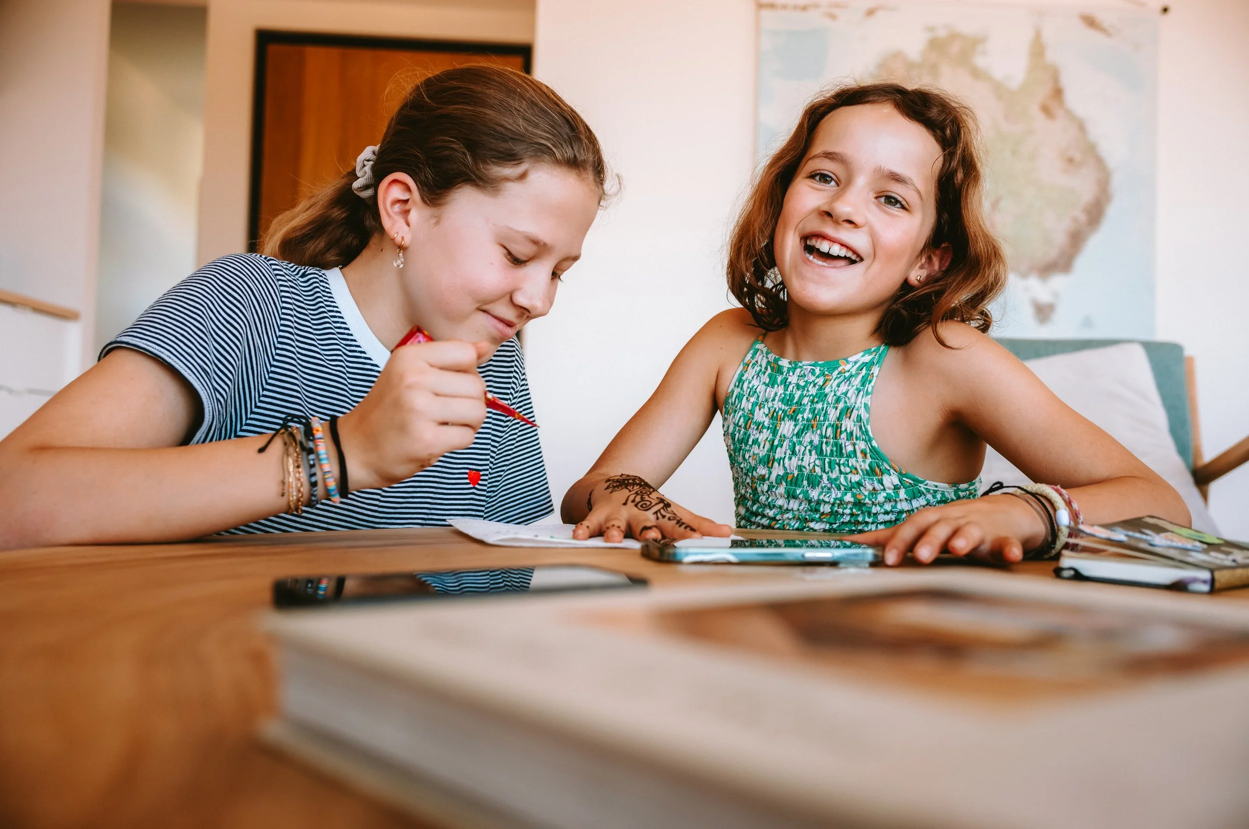 Two young girls sitting at a wooden table, one painting her nails and the other smiling. There are smartphones, books, and a world map on the wall behind them.