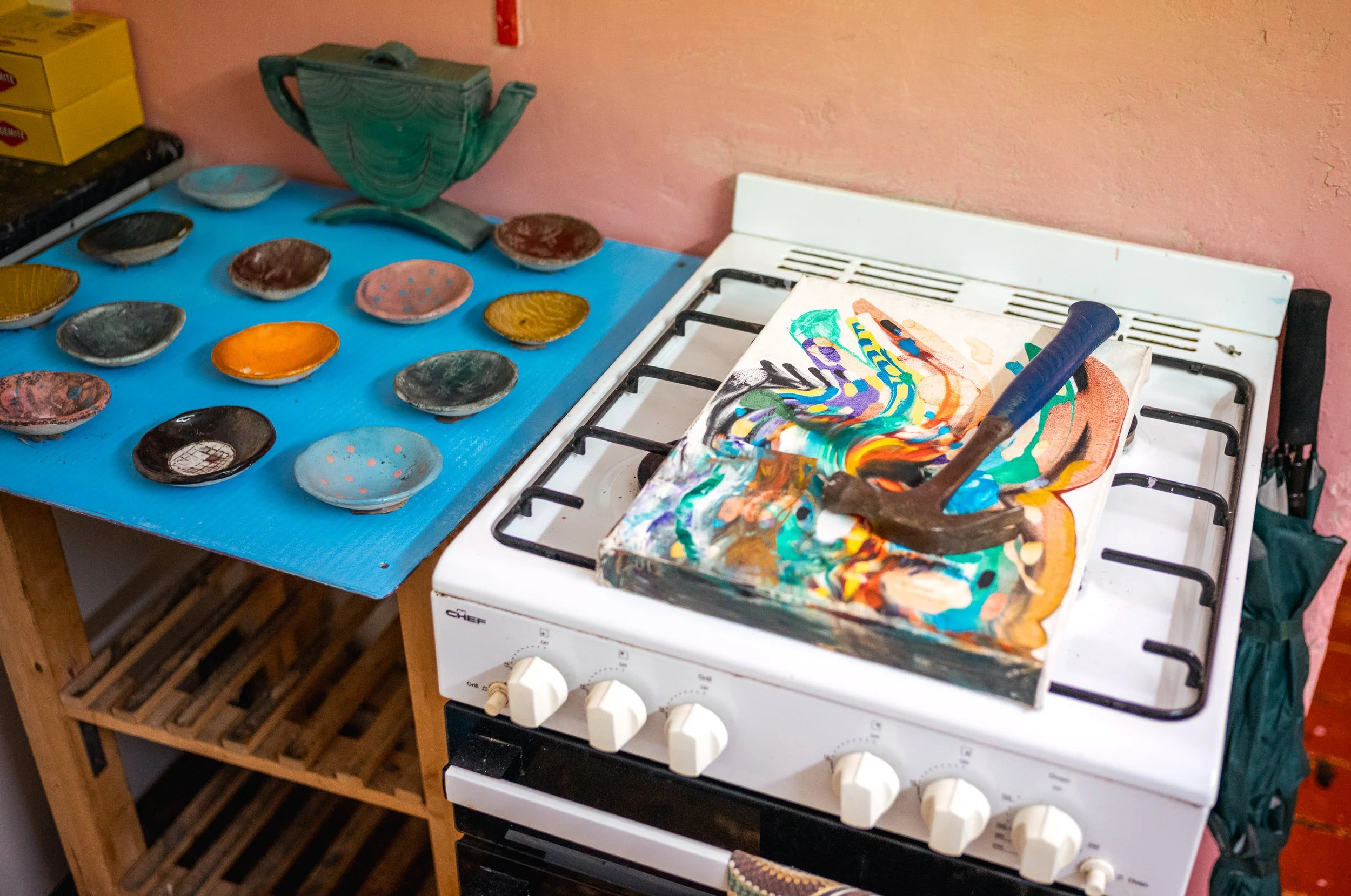 A kitchen stove with a colorful abstract painting on a canvas and a hammer on top. Behind it is a blue tabletop with various small ceramic bowls, some painted in different colors, and a green decorative bowl. The background wall is pink with some vis