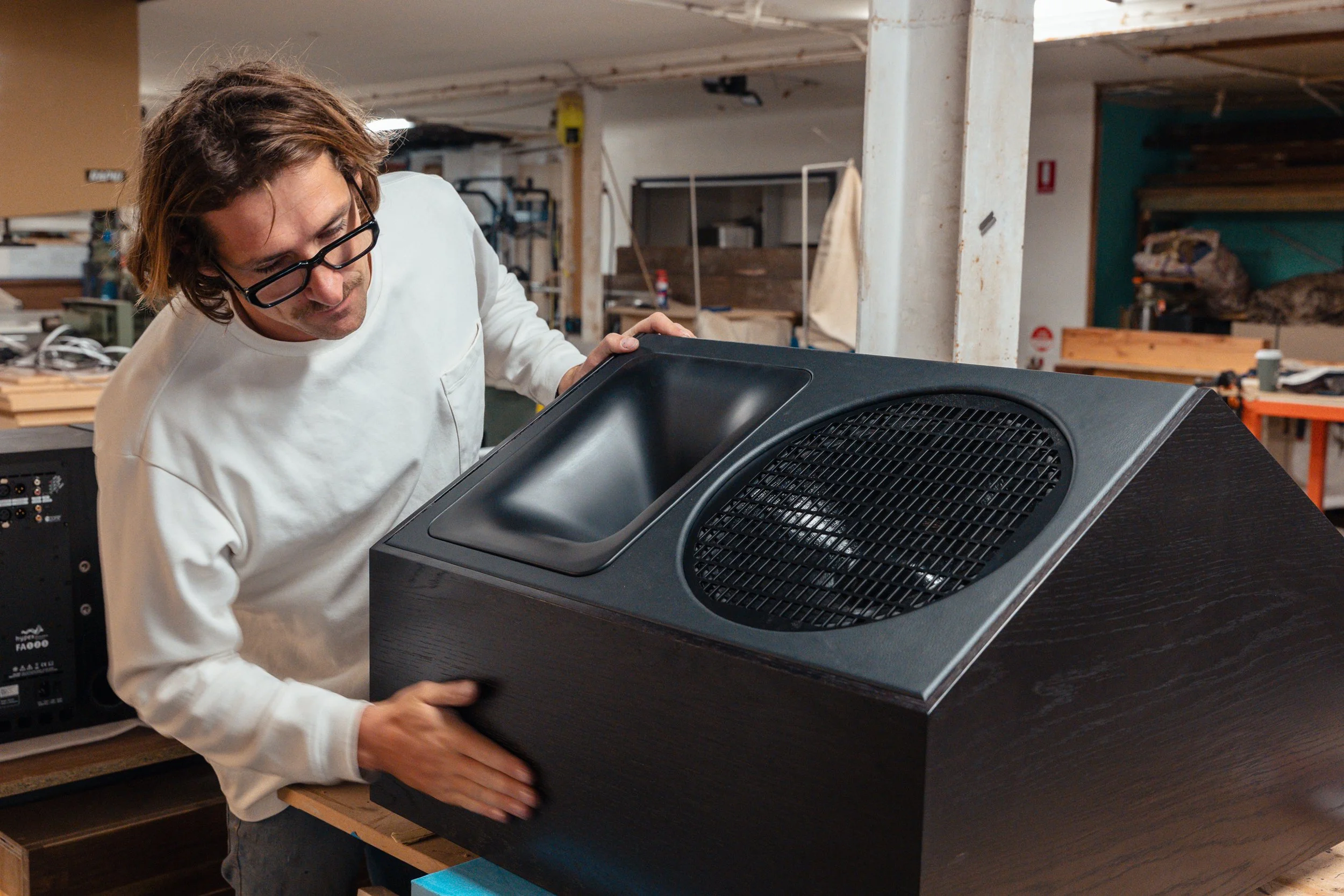 Man inspecting a black, cube-shaped speaker with a large round grill inside a workshop.