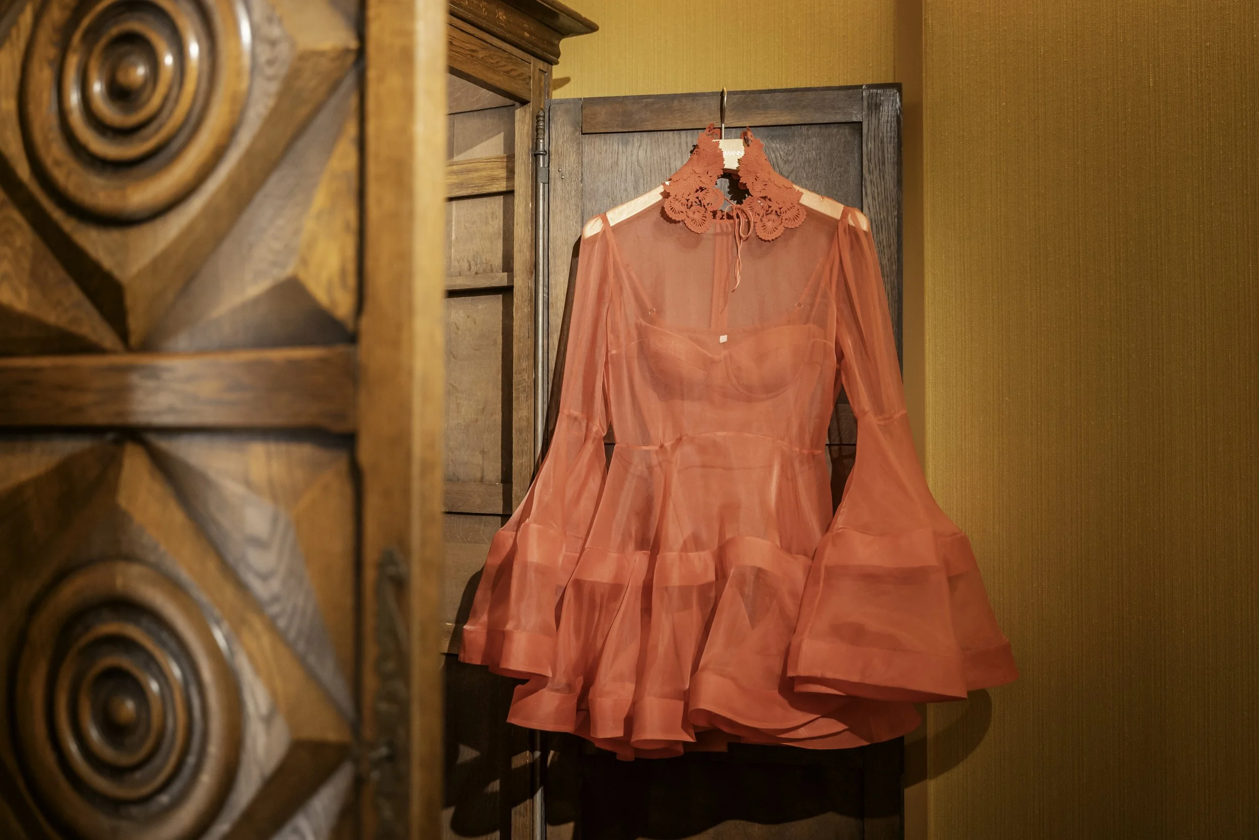 A sheer, red dress with long bell sleeves and floral lace detail around the collar, hanging on a hanger in front of a wooden screen and a warm-toned yellow wall.