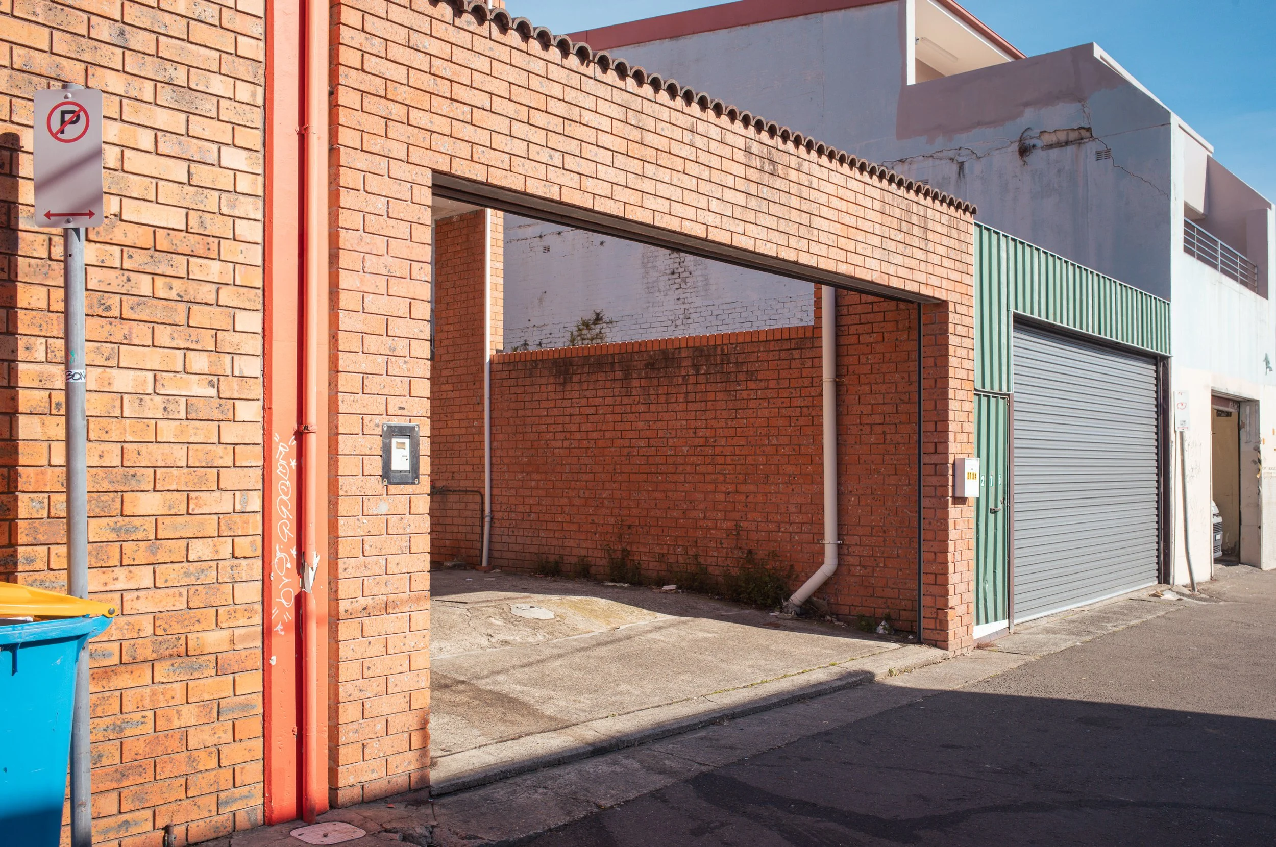 An urban alleyway with brick walls, a yellow and blue trash bin, and a garage door with a gray metal roll-up shutter, under clear blue sky.