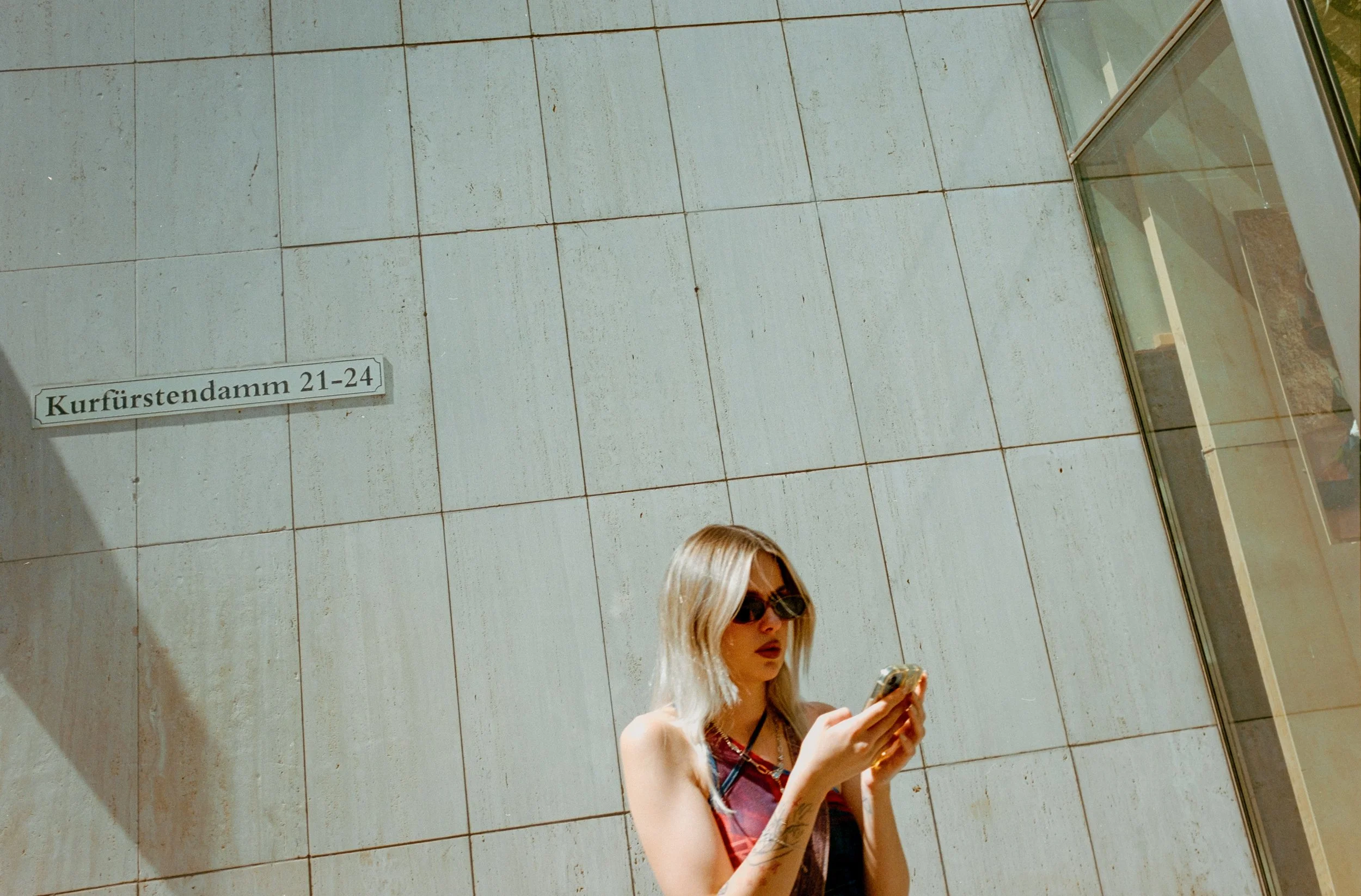 A woman with blonde hair, wearing sunglasses and a sleeveless top, stands against a beige tile wall while looking at her smartphone. There is a street sign on the wall that reads 'Kurfürstendamm 21-24' in the upper left corner.