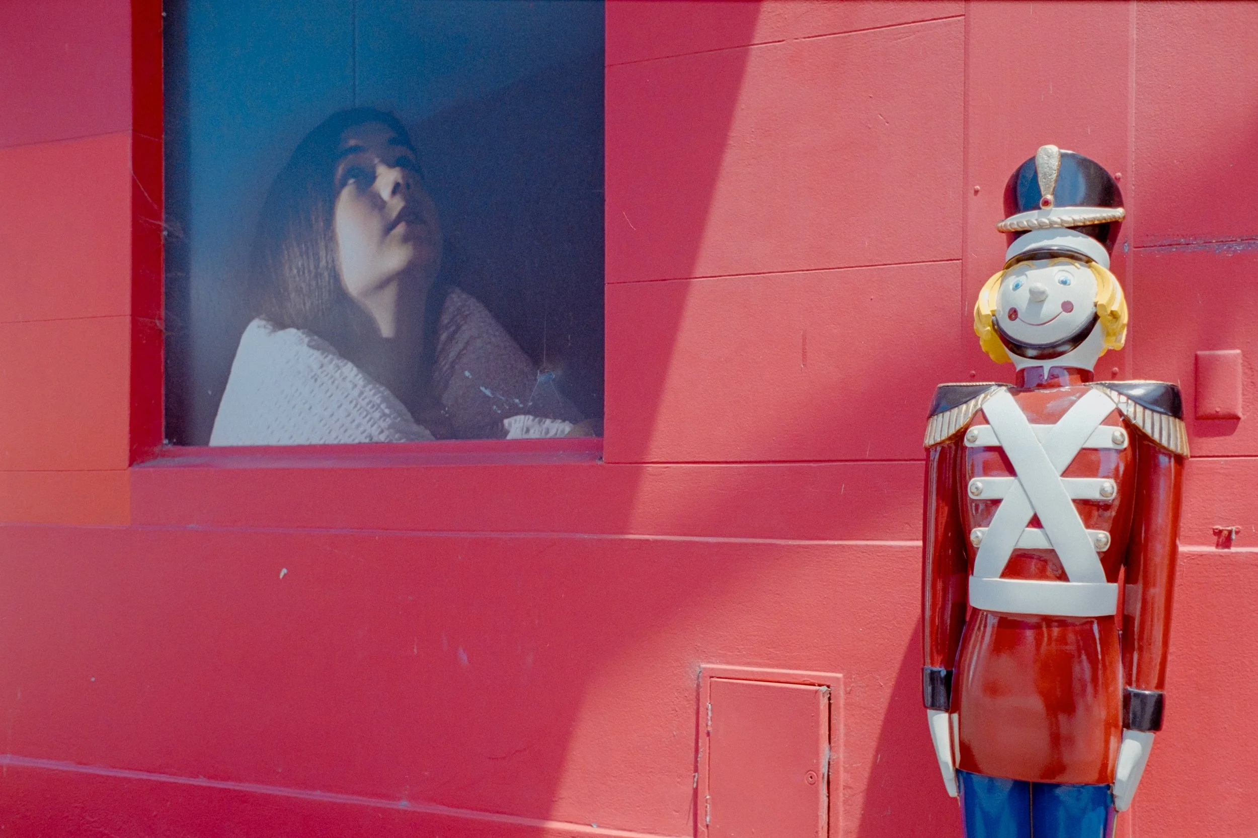 A woman looking out a window in a red building with a toy soldier statue next to the wall.