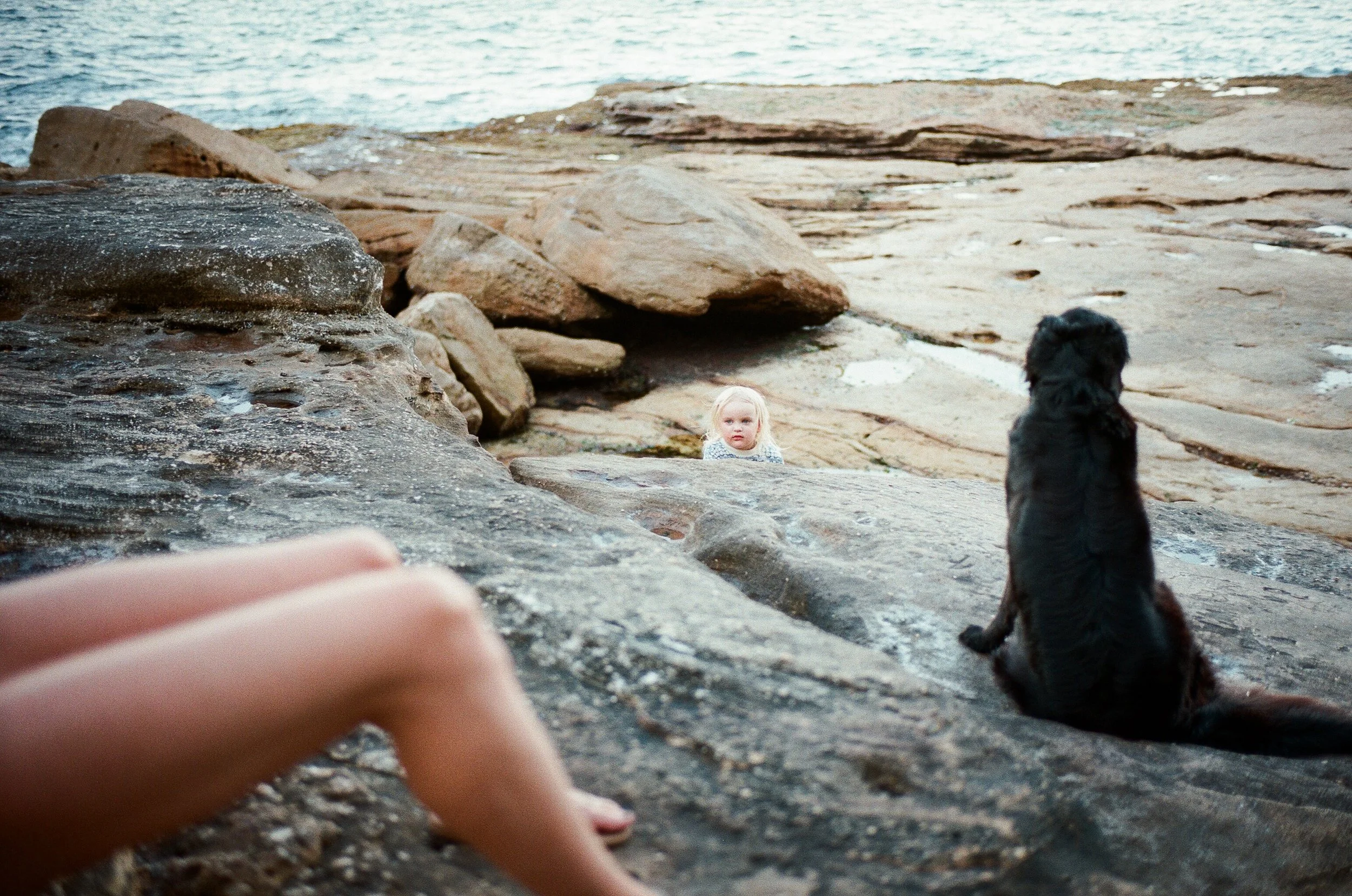 A person relaxing on rocky terrain near the water, with two children in the background, one of whom has blonde hair and the other has dark hair, both sitting on the rocks and facing the person.