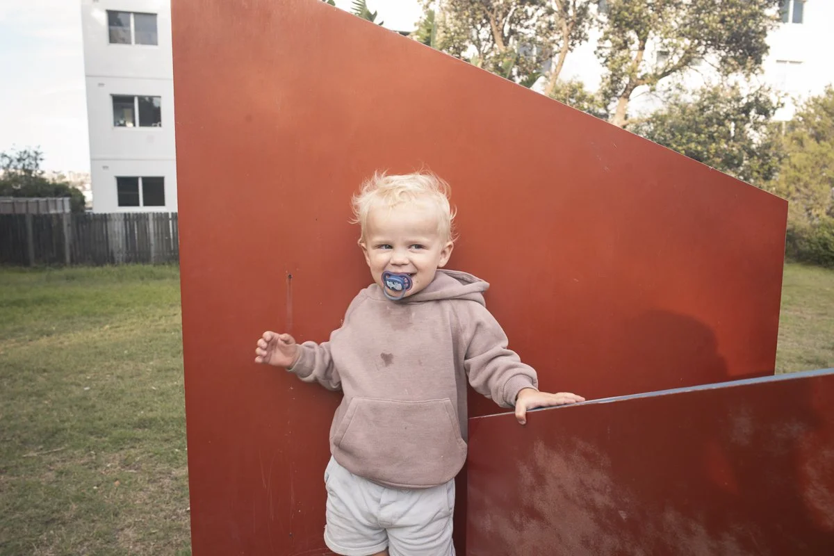 A young child with blonde hair, wearing a beige hoodie and light-colored shorts, standing outdoors next to a large, red geometric sculpture in a grassy area with trees and a building in the background. The child has a pacifier in their mouth and is s
